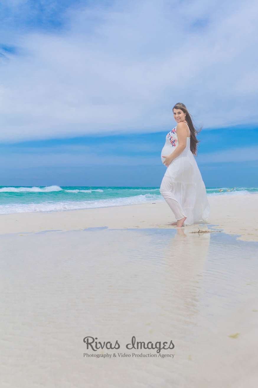 Pregnant woman in a flowing white dress cradling her belly while walking along the shoreline in Seaside, Florida