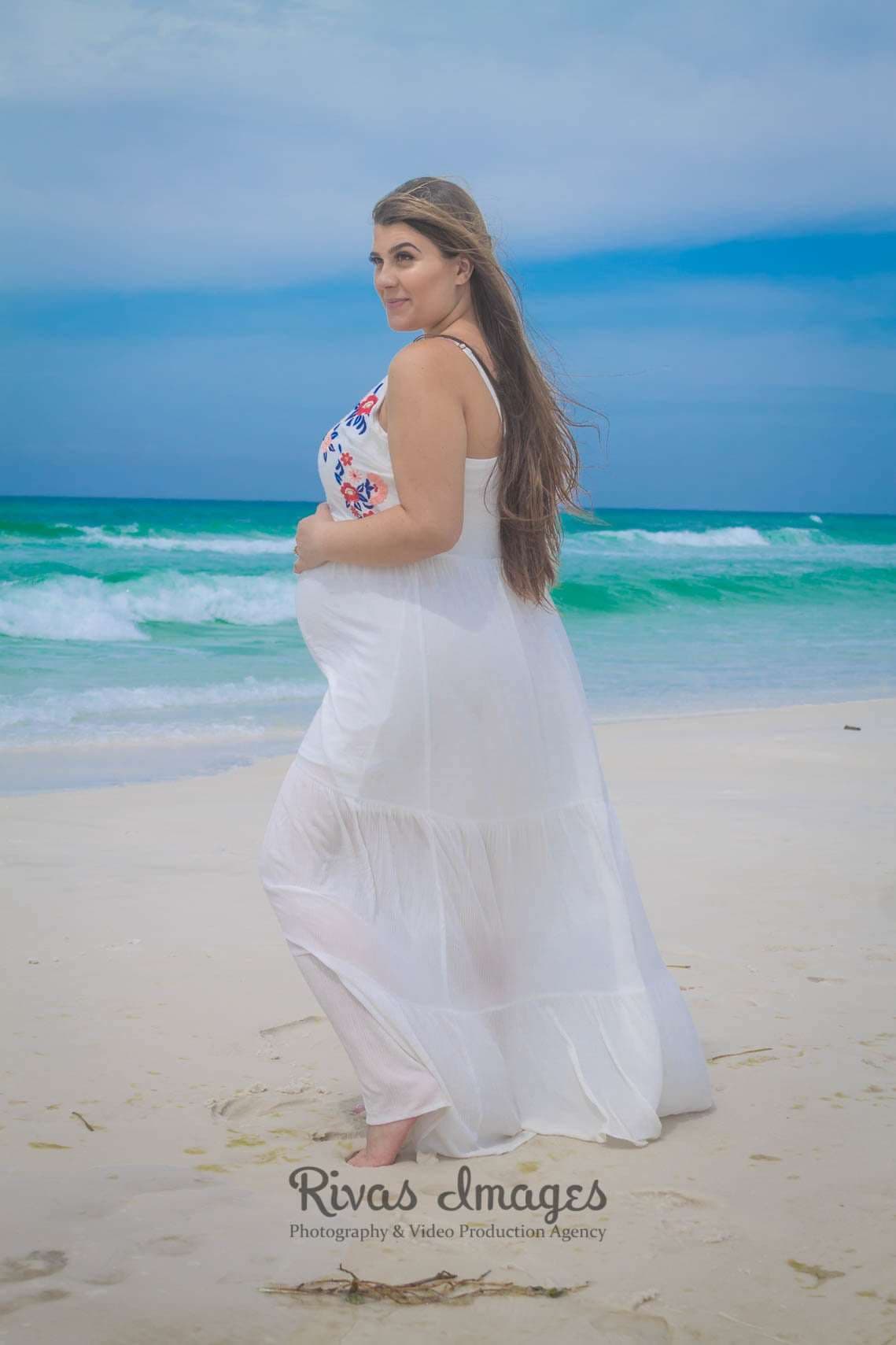 Pregnant woman in a flowing white dress cradling her baby bump on the sand beside turquoise water in Seaside, Florida