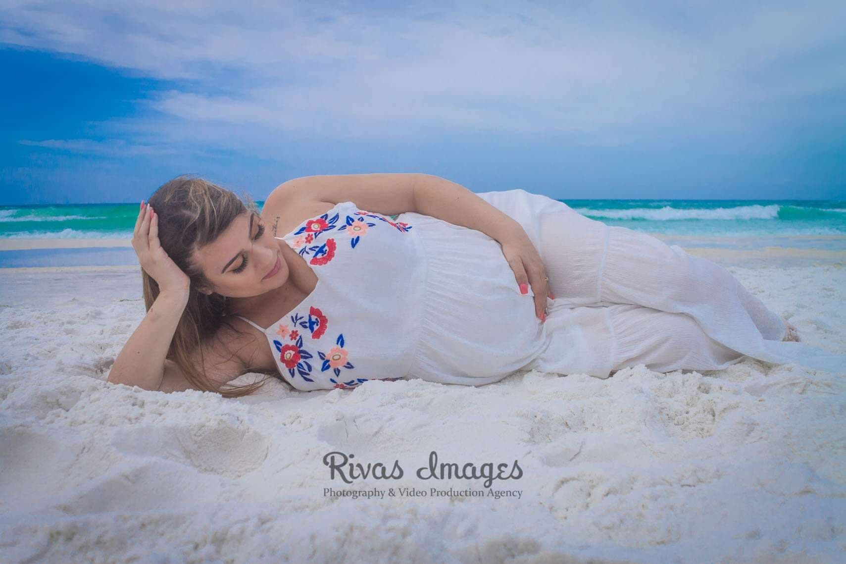 Pregnant woman in a white embroidered dress lying on white sand, resting a hand on her belly by turquoise water