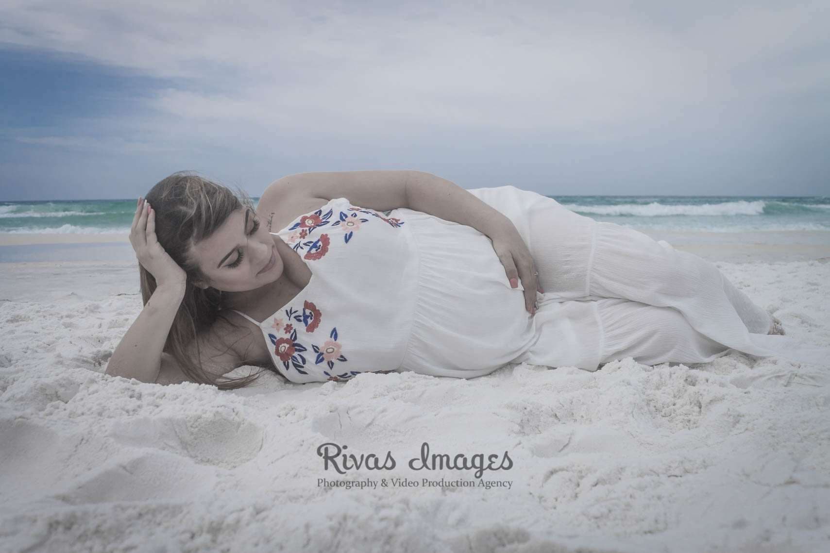 Pregnant woman in a white embroidered dress reclining on white sand, hand on her belly, with the ocean behind her