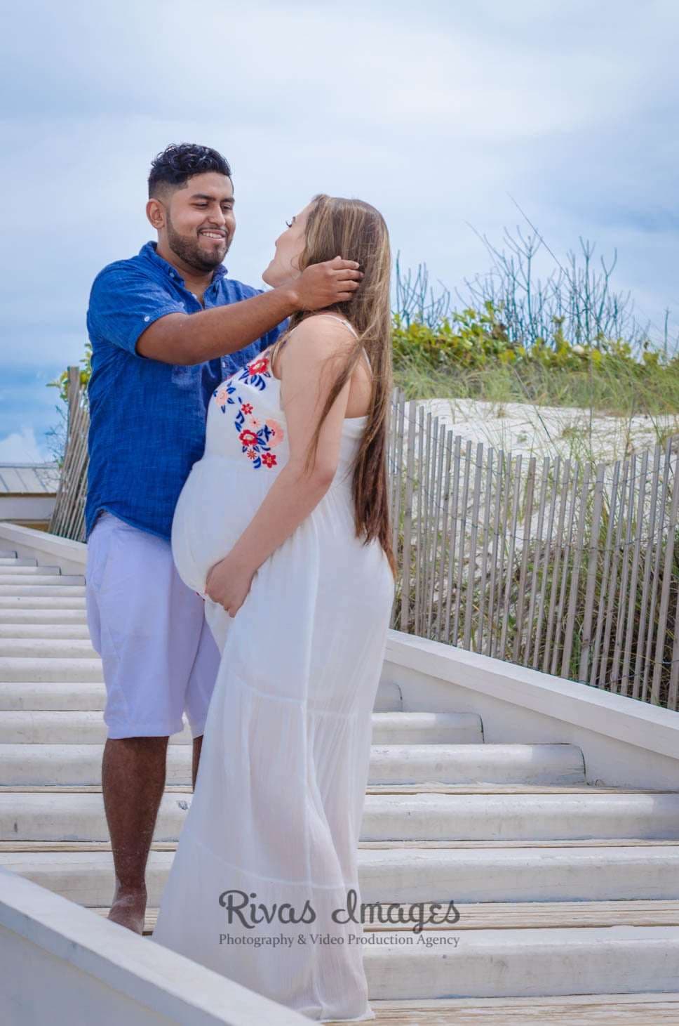 Expectant couple embracing on white beach steps, man in blue shirt tucking her hair behind her ear, in Seaside, Florida