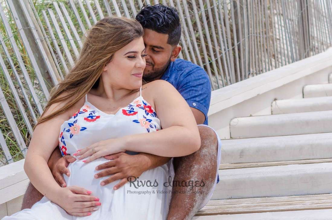 Expectant couple embracing on wooden beach steps, her hands on her baby bump in a white floral dress