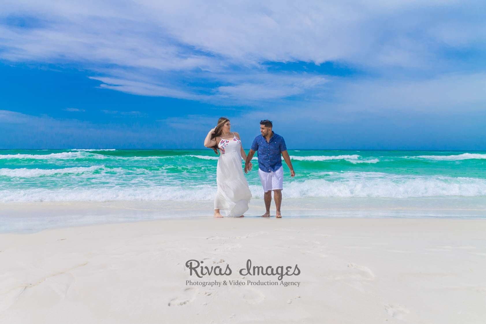 Couple holding hands and walking along white sand toward the camera beside turquoise waves under a bright blue sky