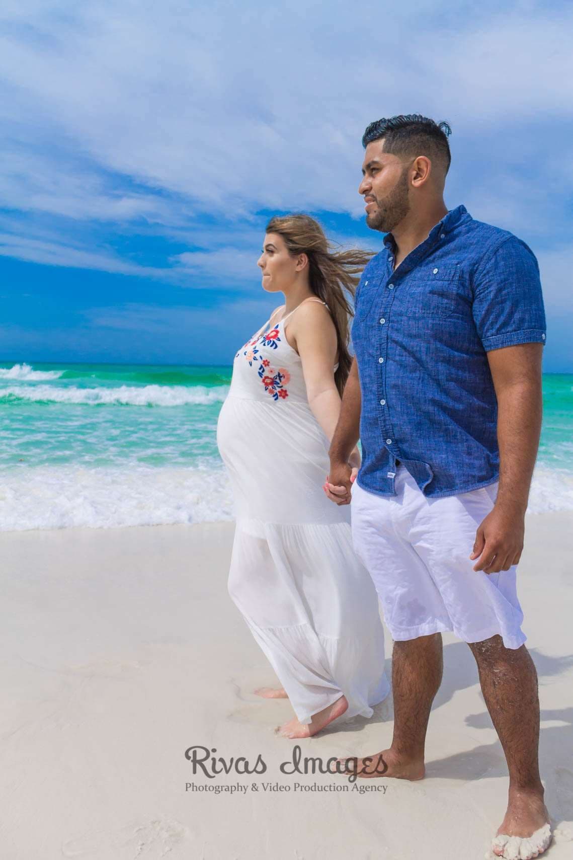 Expectant couple holding hands and walking barefoot on white sand beside turquoise water in Seaside, Florida
