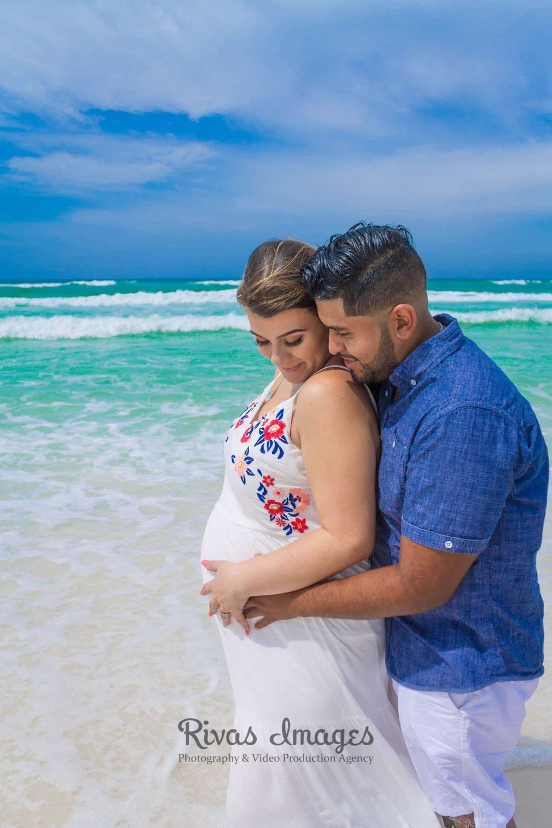 Expectant couple embracing on the beach in Seaside, Florida, his hands on her baby bump, turquoise waves behind them