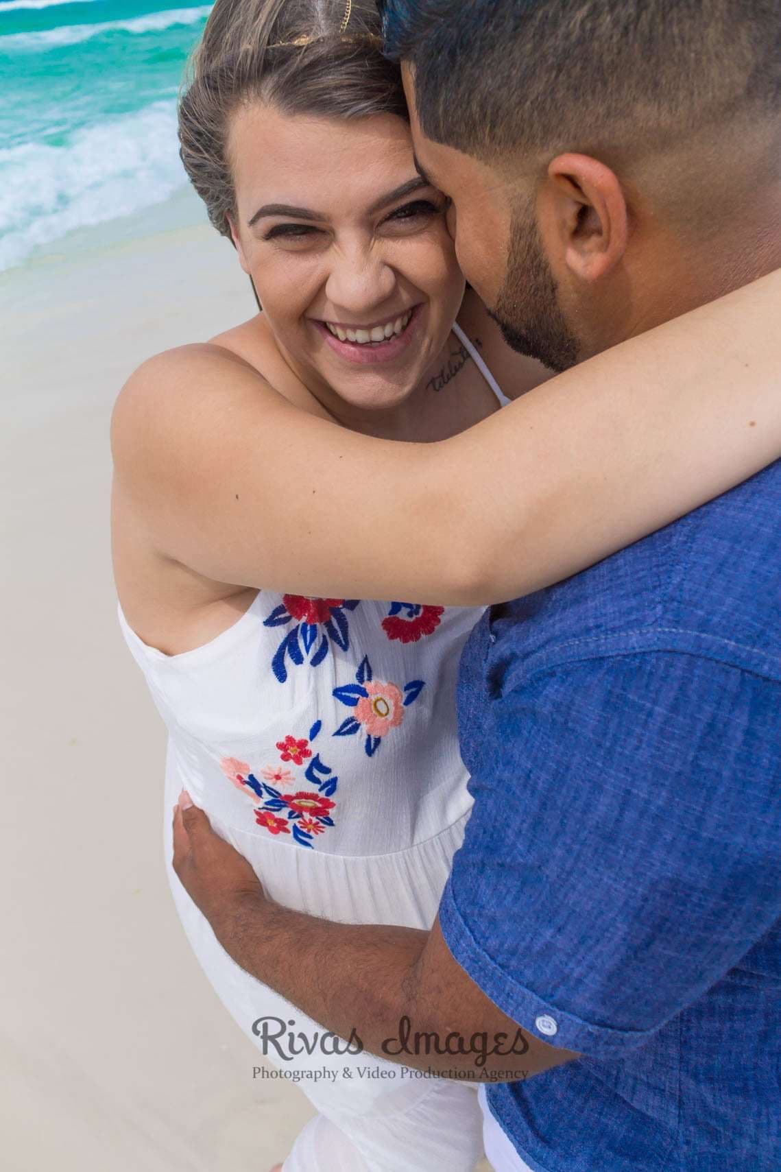 Laughing woman in a white floral sundress embraced by a man in a blue shirt on a beach by turquoise water