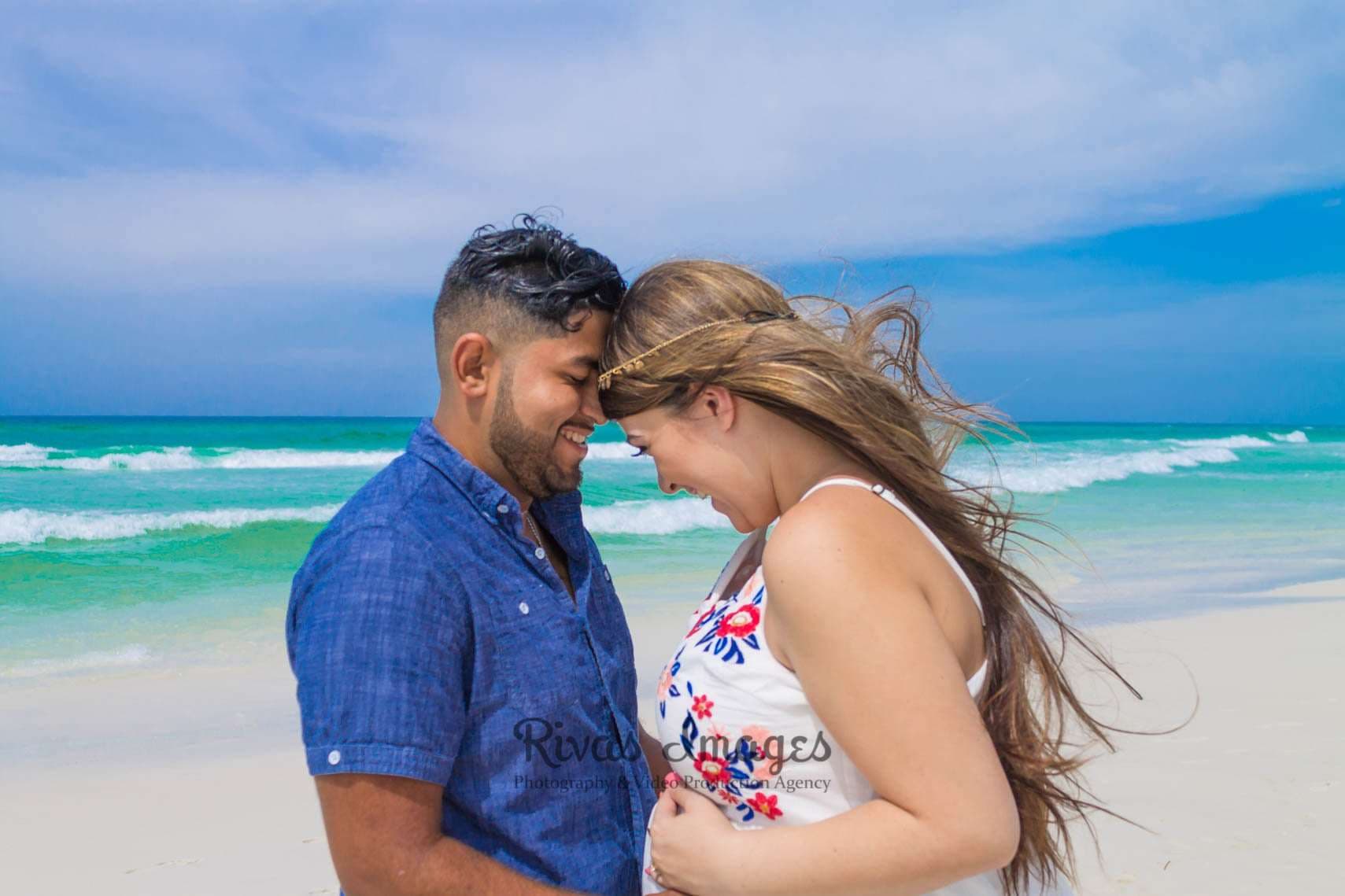 Smiling couple touching foreheads on a white sand beach as the woman's hair blows, turquoise waves behind them