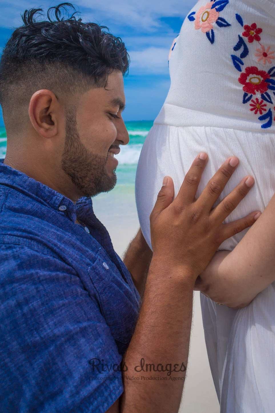 Smiling man in a blue shirt rests his hand on a pregnant woman's belly on a Seaside beach with turquoise water