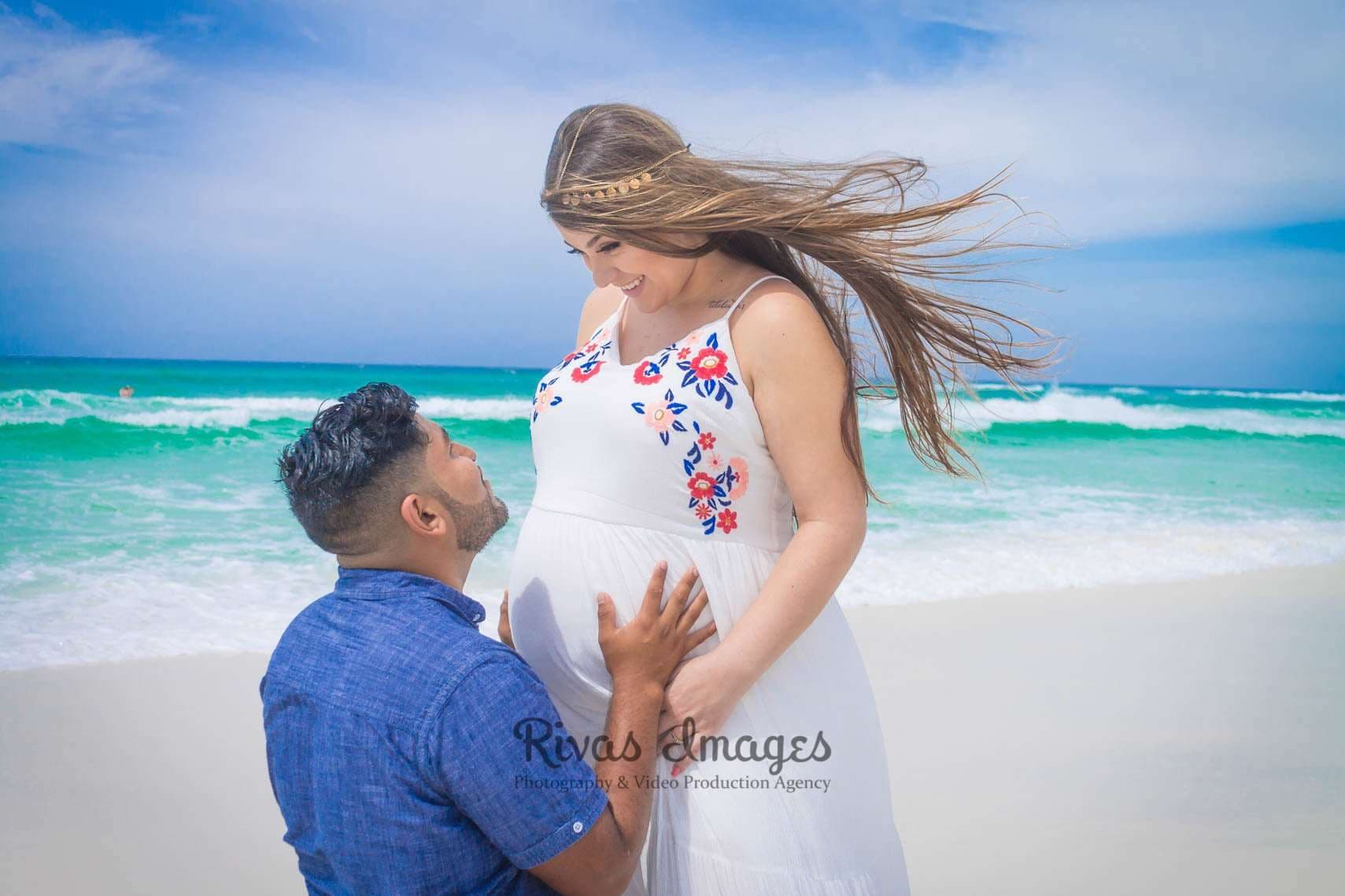 Man kneeling in the sand with hands on his pregnant partner's belly as she smiles down, hair blowing by turquoise water