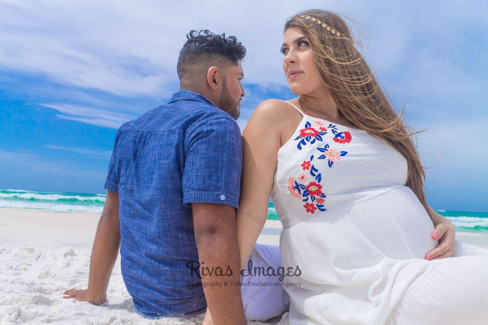Expectant couple sitting close on a white sand beach, the woman in a white embroidered dress looking back at him