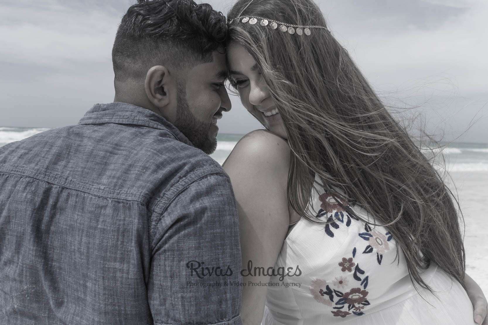 Black-and-white shot of a couple touching foreheads and smiling on a windy beach, woman in embroidered dress and headpiece