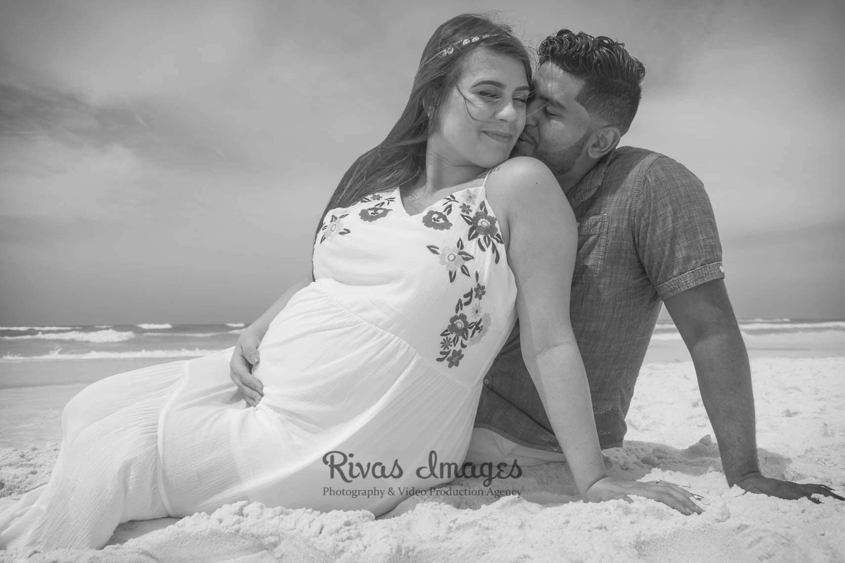 Black-and-white maternity portrait of a man kissing his partner's shoulder as she sits on the beach holding her belly