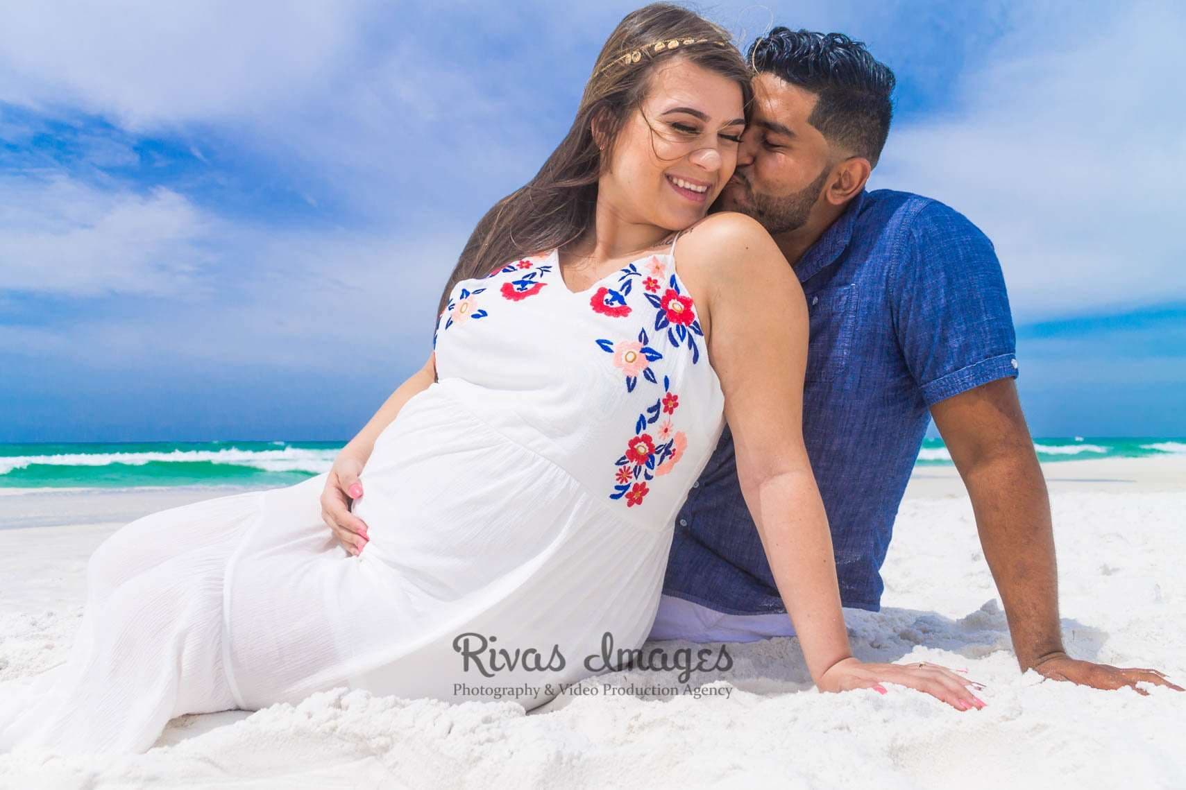 Smiling pregnant woman in a white embroidered dress cradles her belly as her partner kisses her cheek on a white sand beach