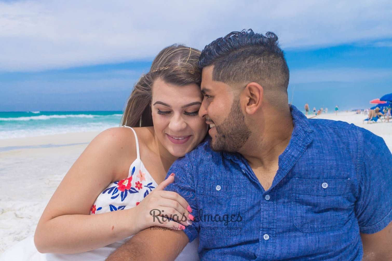 Smiling couple sitting close on a white sand beach, her head resting on his shoulder, turquoise water behind them