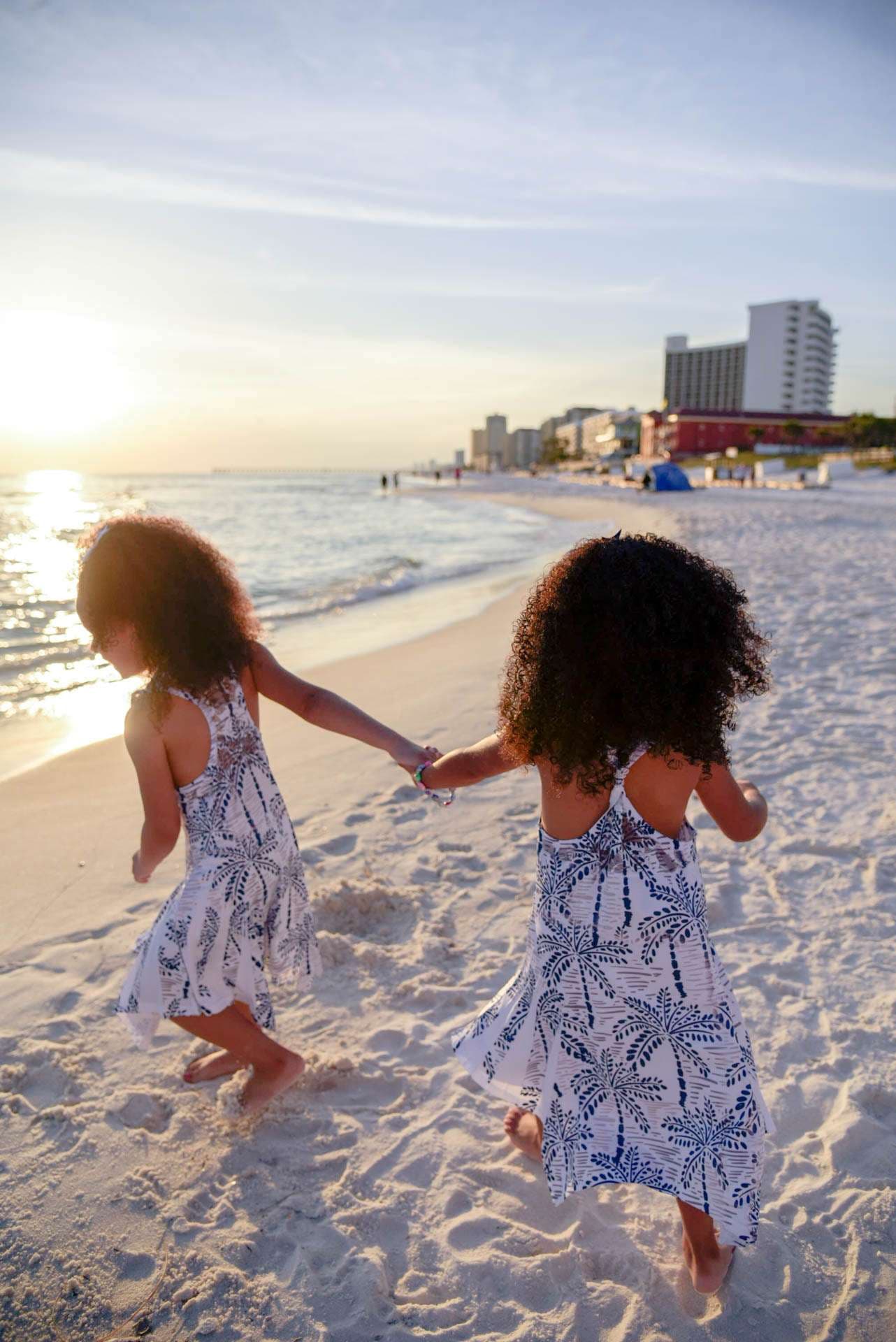 Two girls in matching blue and white palm print dresses holding hands on a sandy beach at golden hour