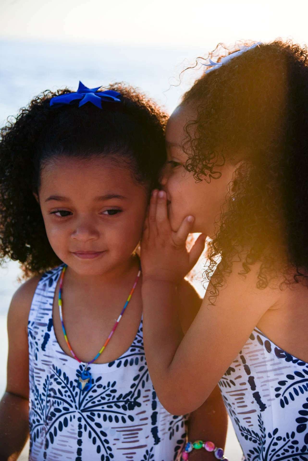 Two young girls in matching palm-print dresses share a whispered secret in warm golden light by the water