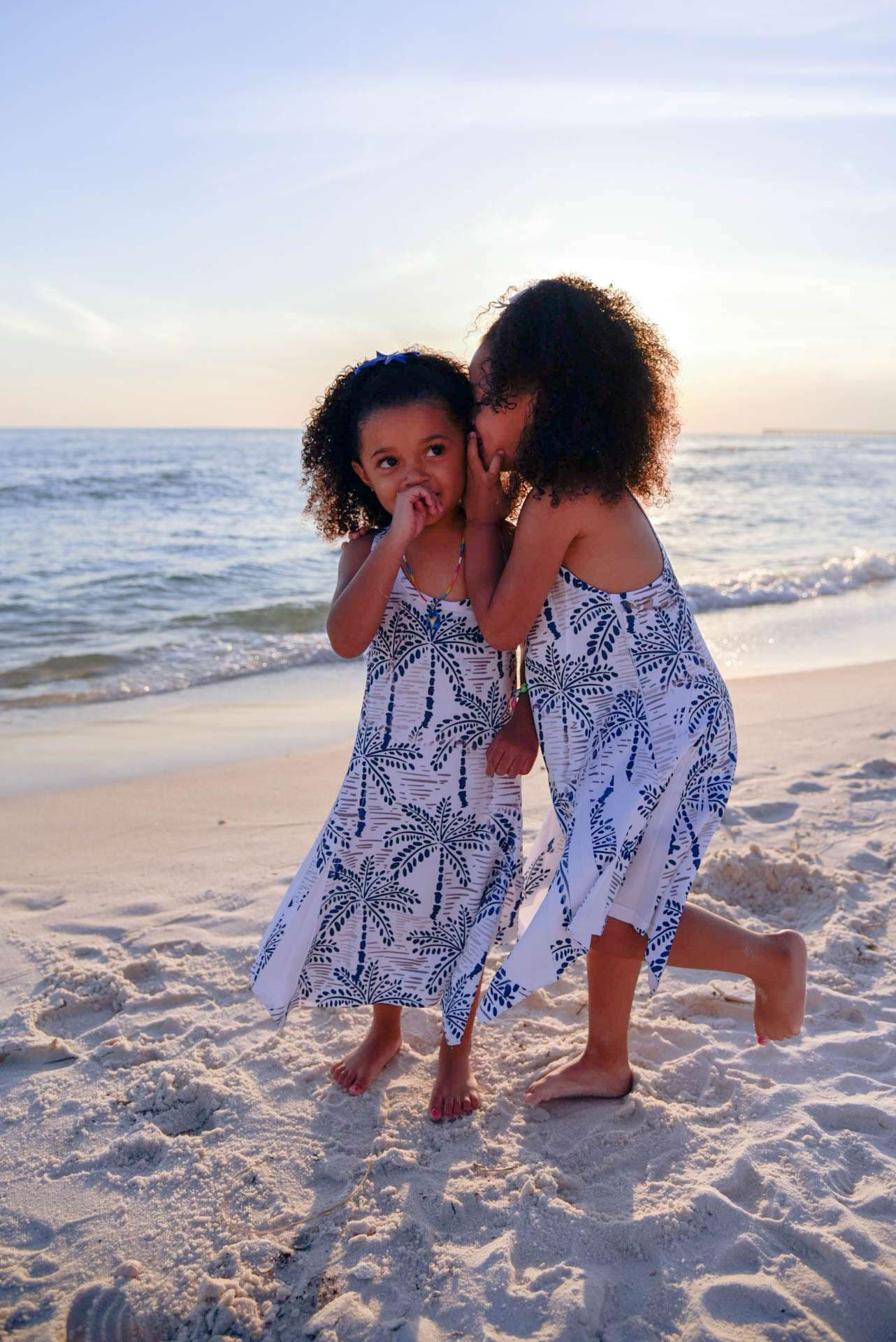 Two girls in matching palm-print dresses whispering secrets on the beach at golden hour, waves behind them