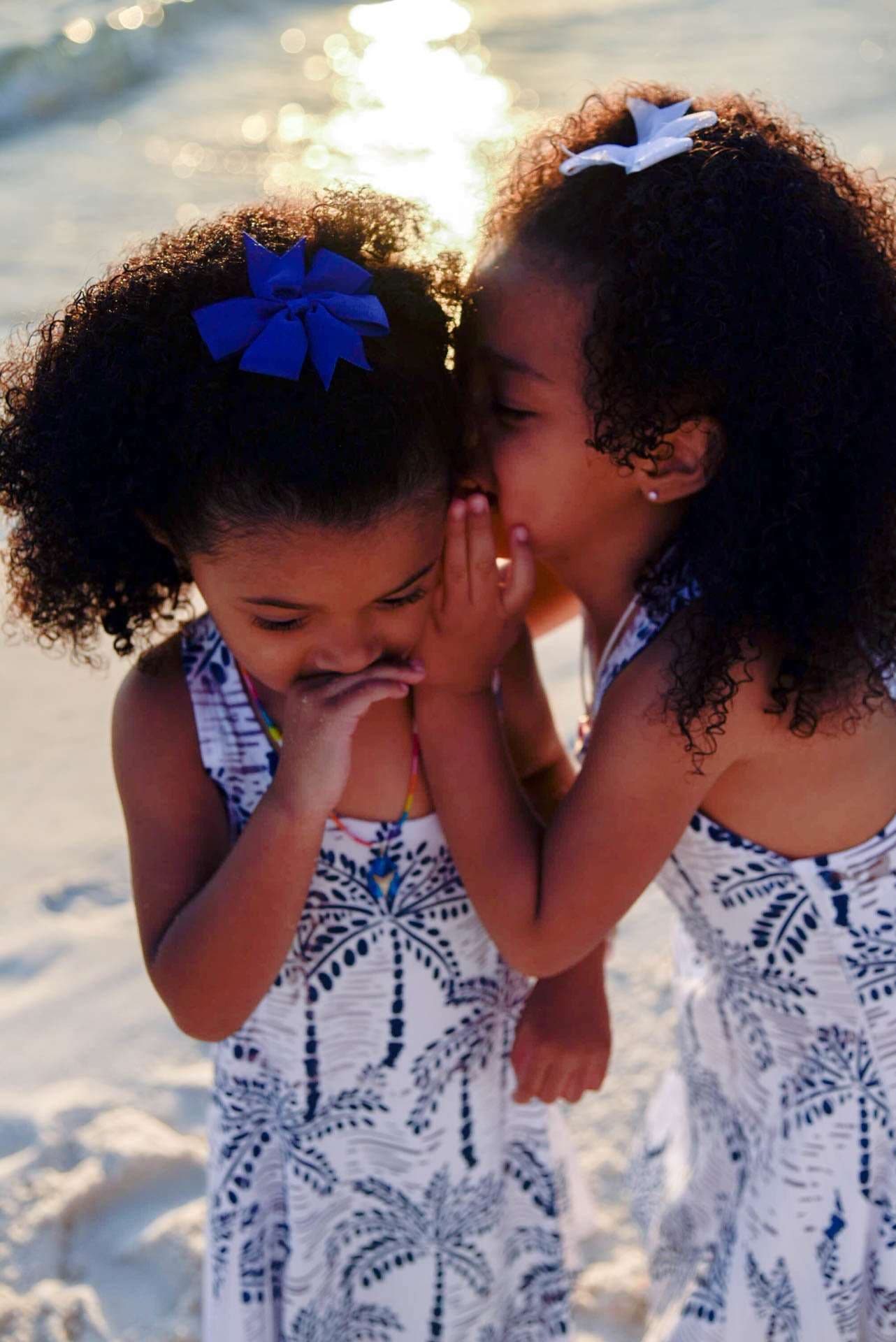 Two girls in matching palm-print dresses whispering a secret on the beach at golden hour, one covering her mouth