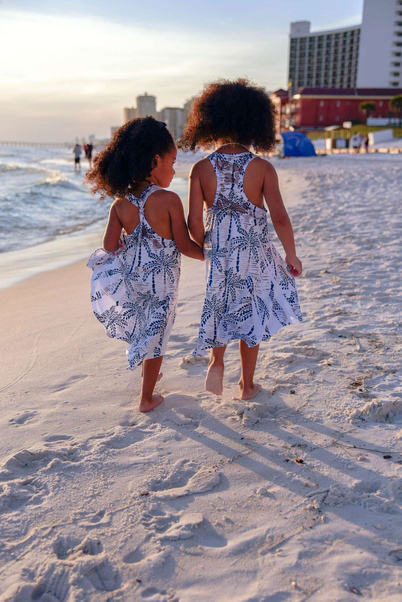 Two young girls in matching palm-print dresses walk barefoot hand in hand along the sandy shore at golden hour