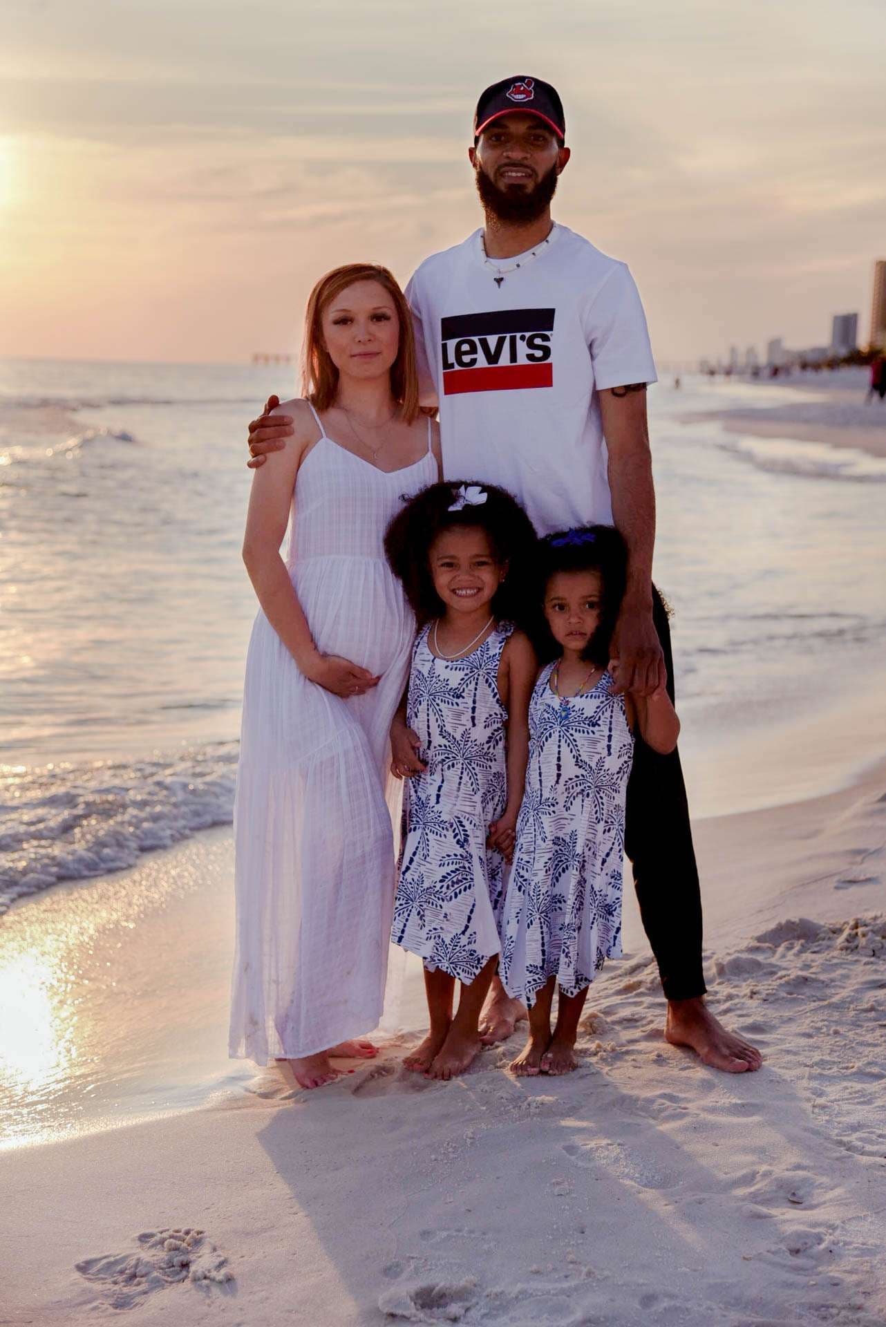 Family of four in white and blue outfits standing barefoot on the beach at golden hour, two young girls in front