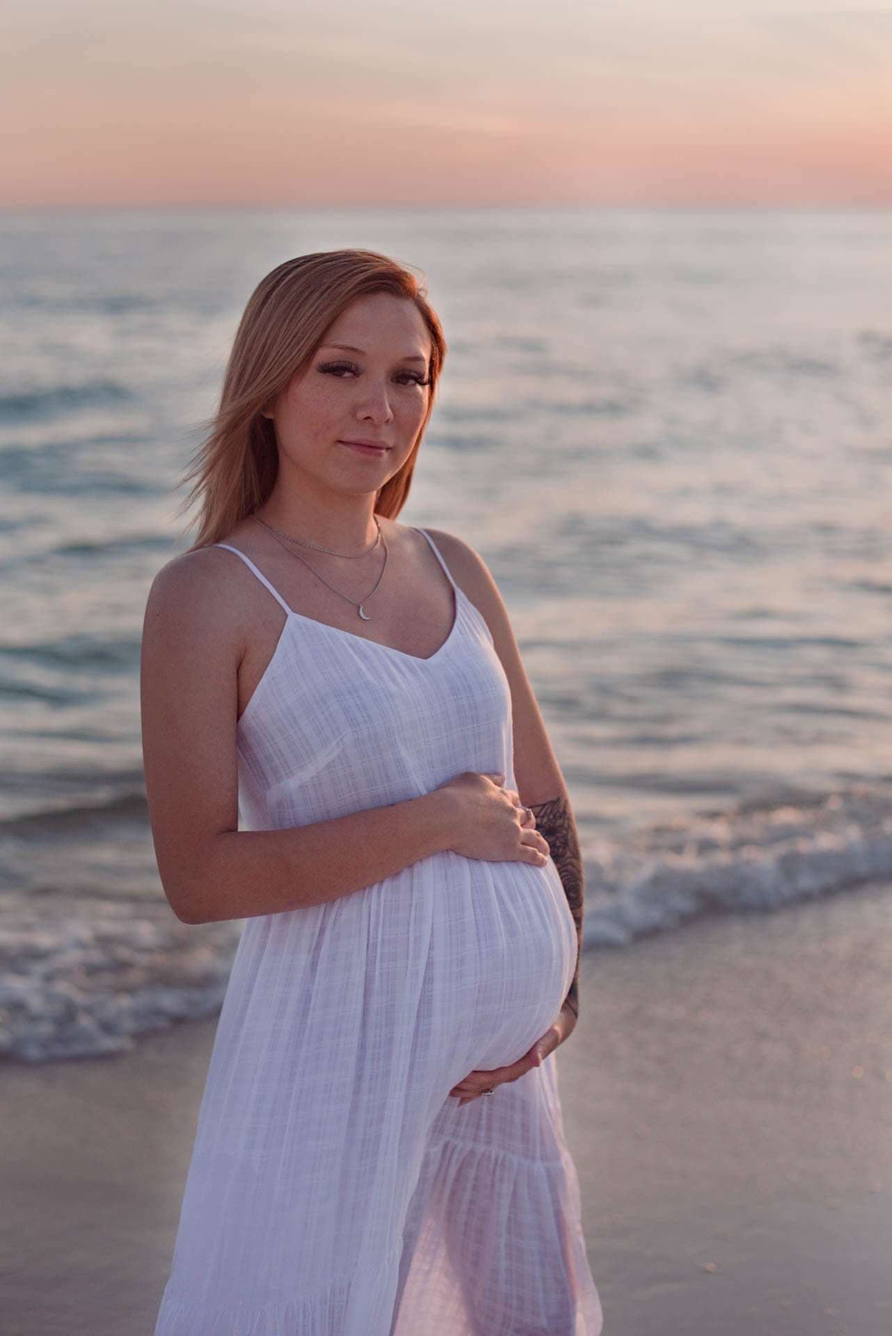 Pregnant woman in a white sundress cradling her baby bump on the beach at sunset