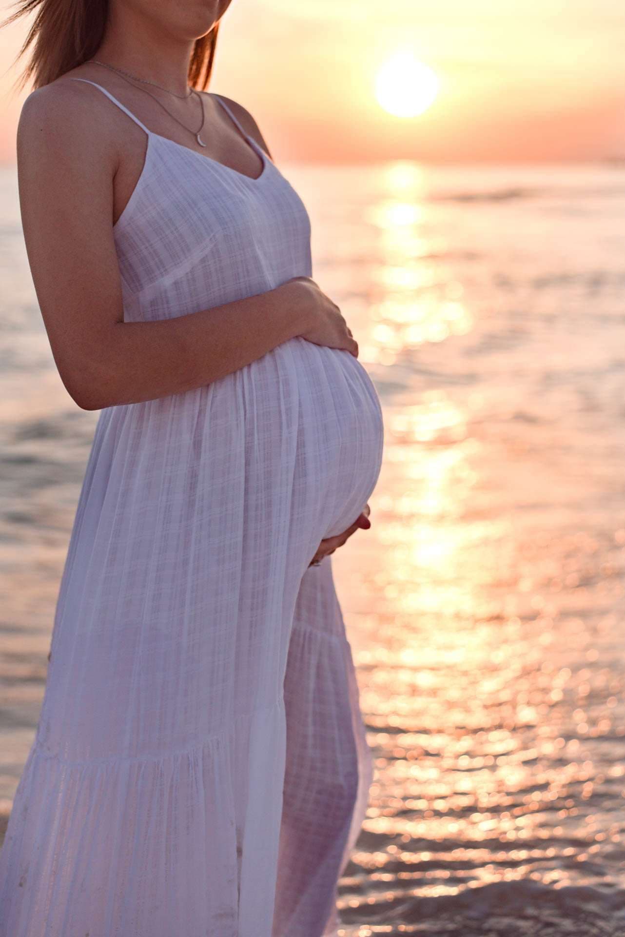 Pregnant woman in a white sundress cradling her baby bump by the water at sunset