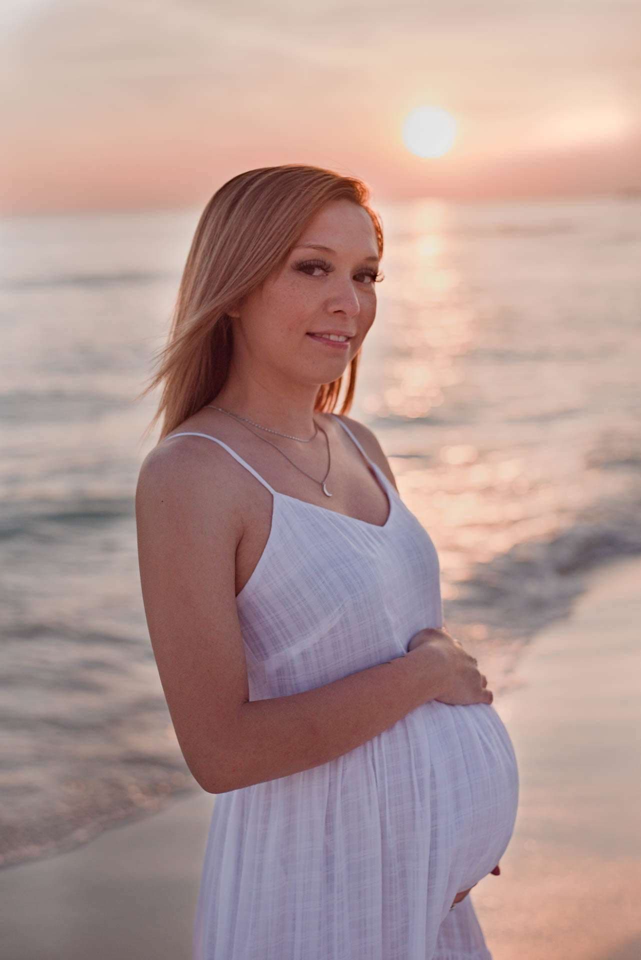 Pregnant woman in a white sundress cradling her baby bump on the shore as the sun sets over the water