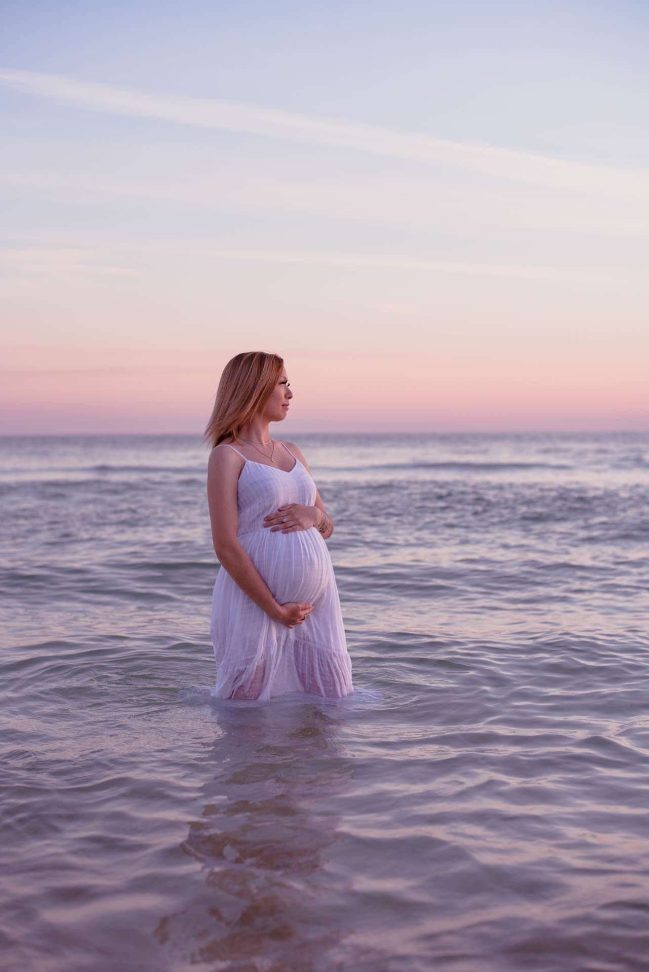 Pregnant woman in a flowing white dress standing in shallow water, cradling her belly under a pastel pink sunset sky