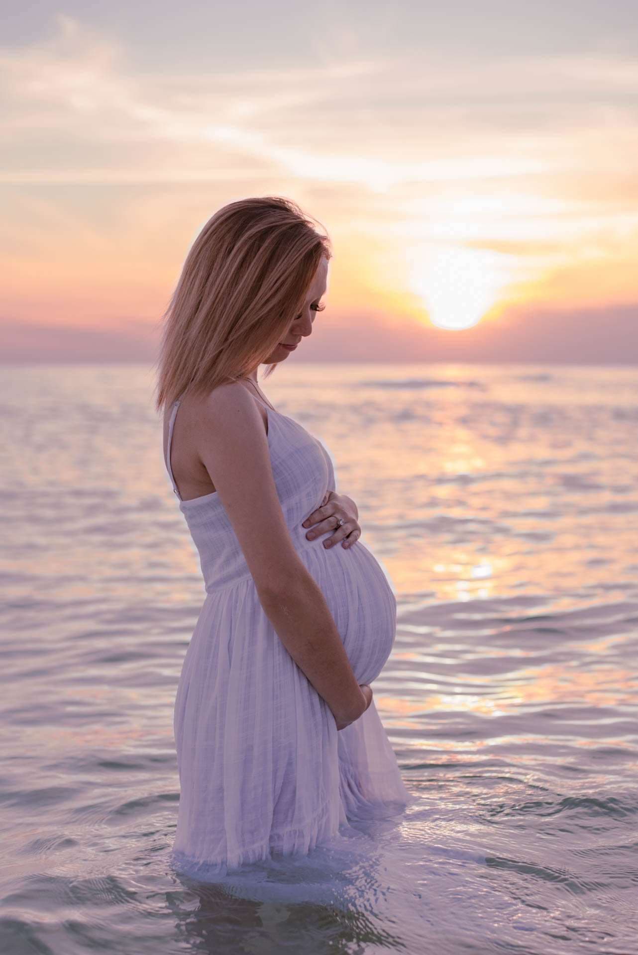 Pregnant woman in a white dress standing in shallow water, cradling her baby bump as the sun sets