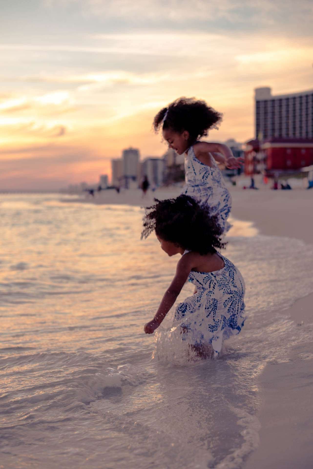 Two young girls in blue and white floral dresses splashing in shallow ocean water at sunset