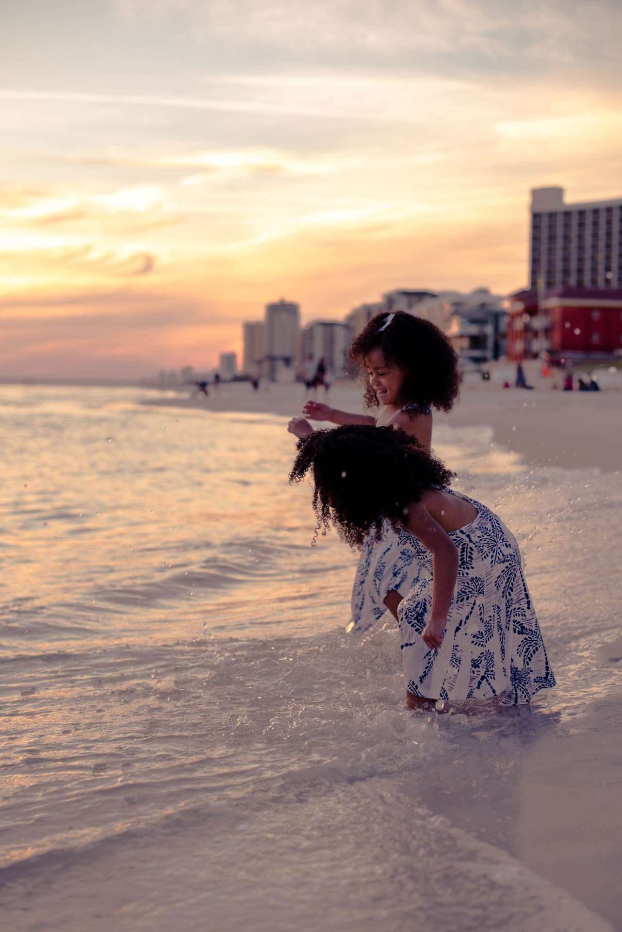 Two children in matching blue and white dresses playing in shallow waves on the beach at golden hour sunset