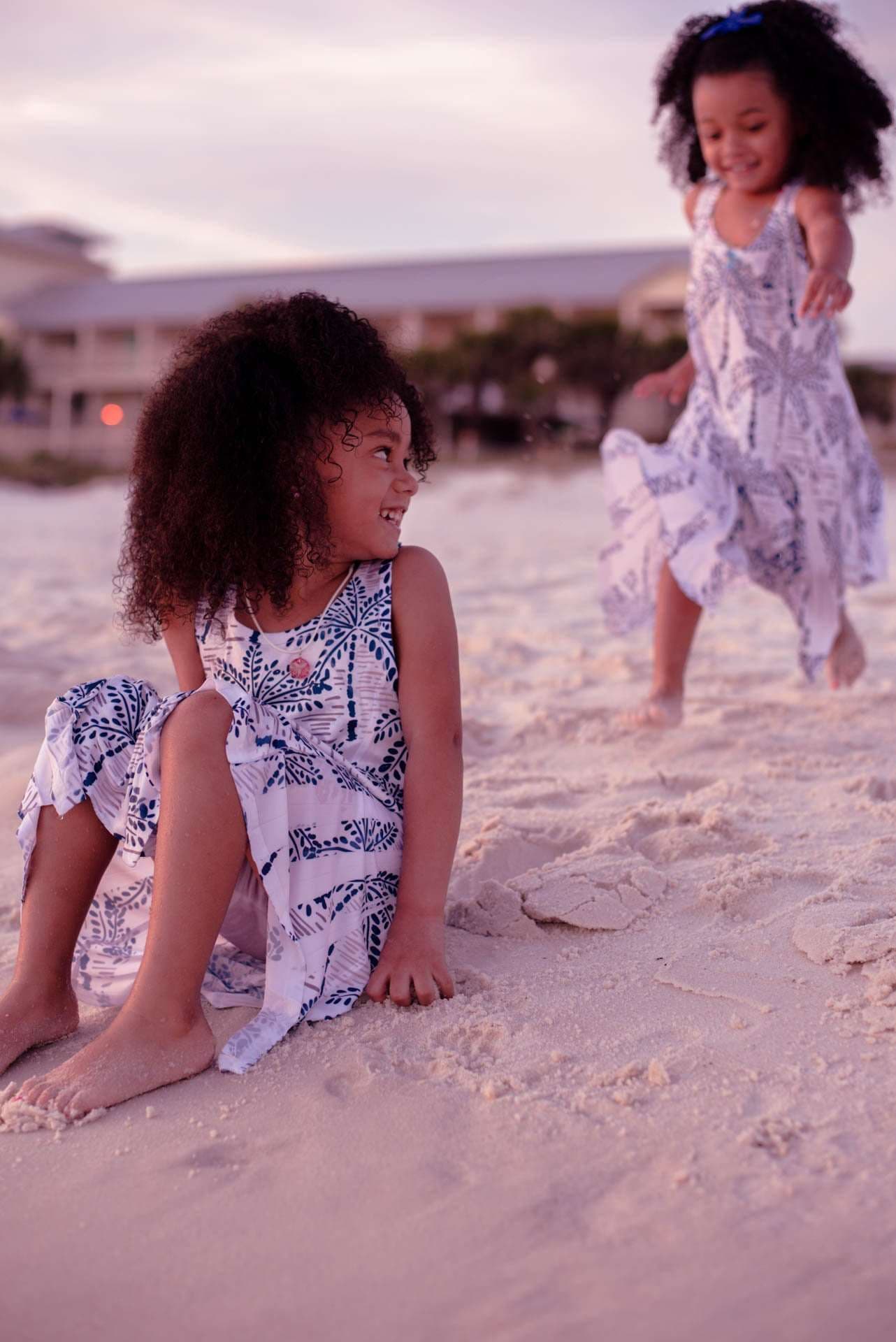 Two girls in matching blue and white palm-print dresses playing on the sand at golden hour, one smiling over her shoulder