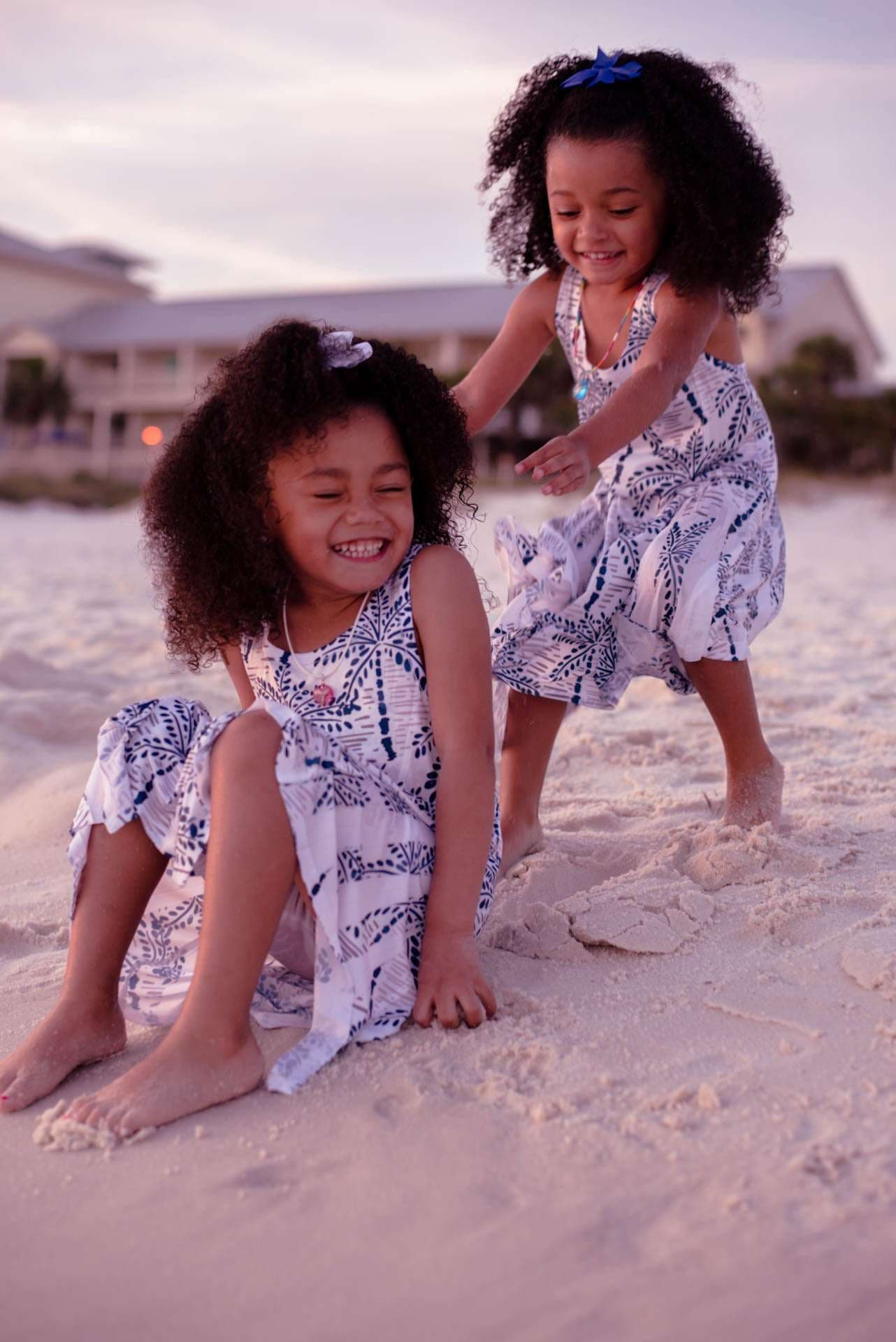 Two smiling girls in matching blue and white dresses playing in the sand at sunset, one seated and one leaning over her