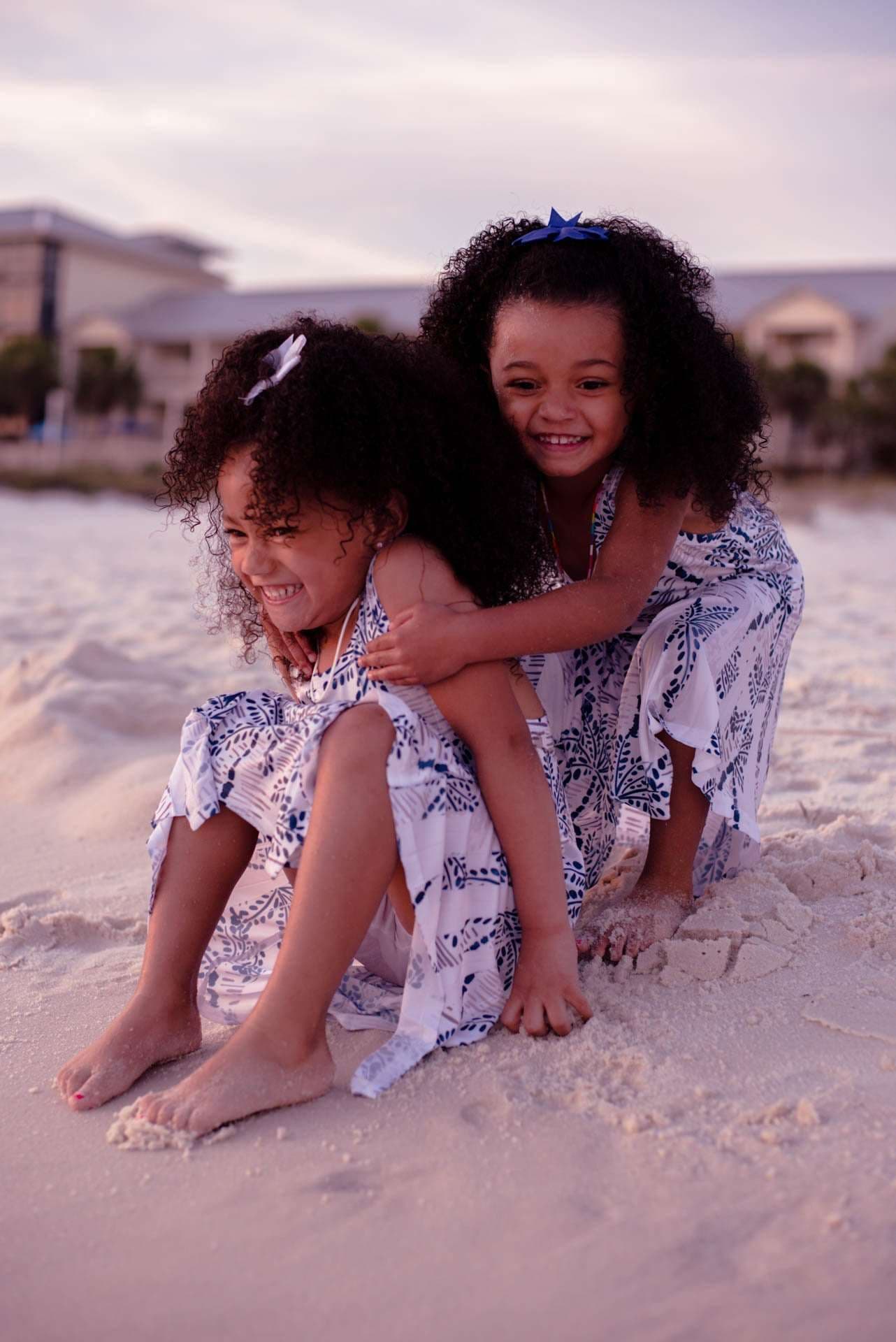 Two smiling girls in matching blue and white dresses hugging and playing in the sand at dusk