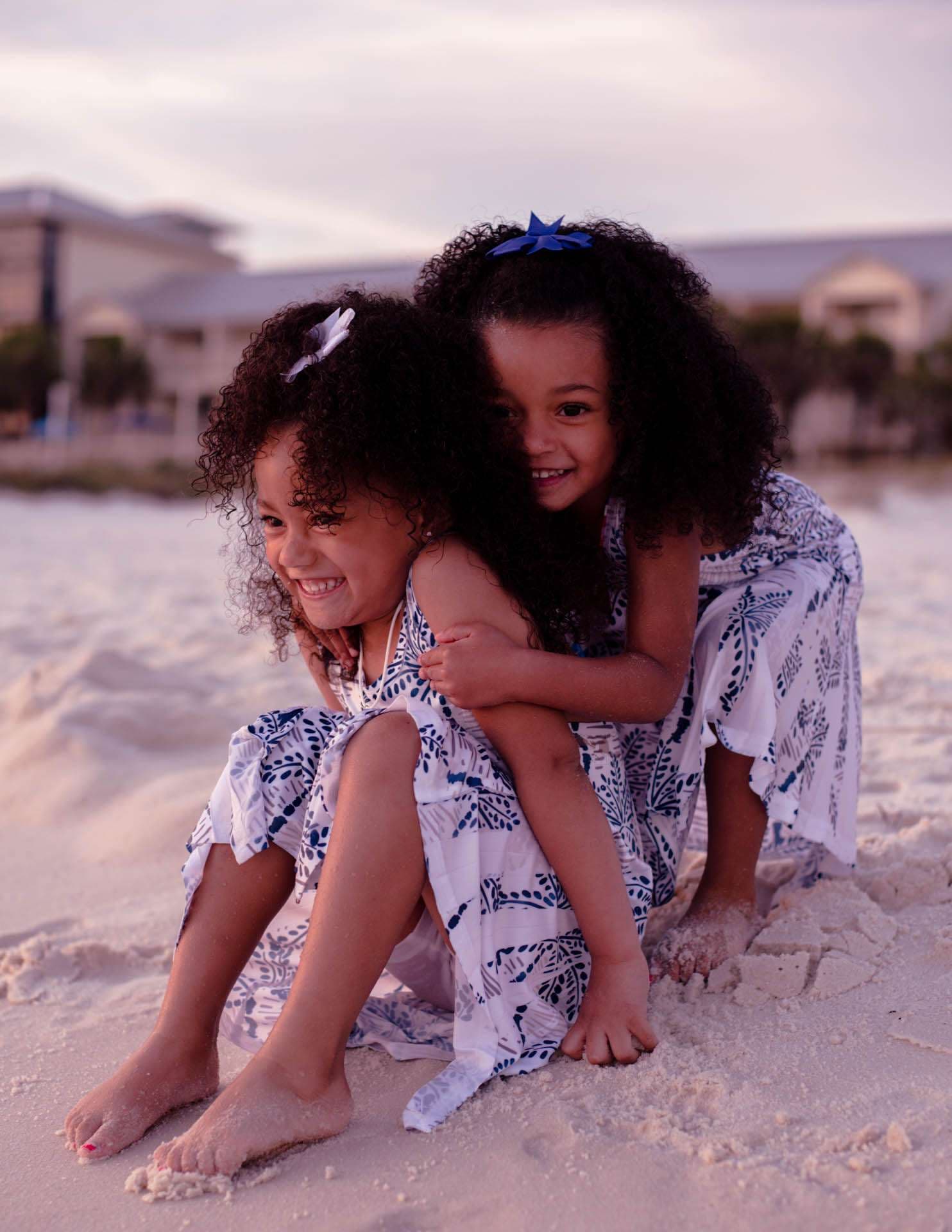 Two smiling girls in matching blue and white dresses hugging on the sand at dusk, one embracing the other from behind