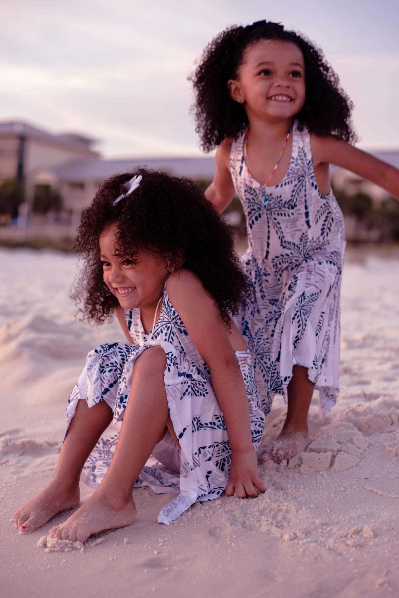 Two smiling girls with curly hair in matching blue and white dresses playing barefoot on the sand at golden hour