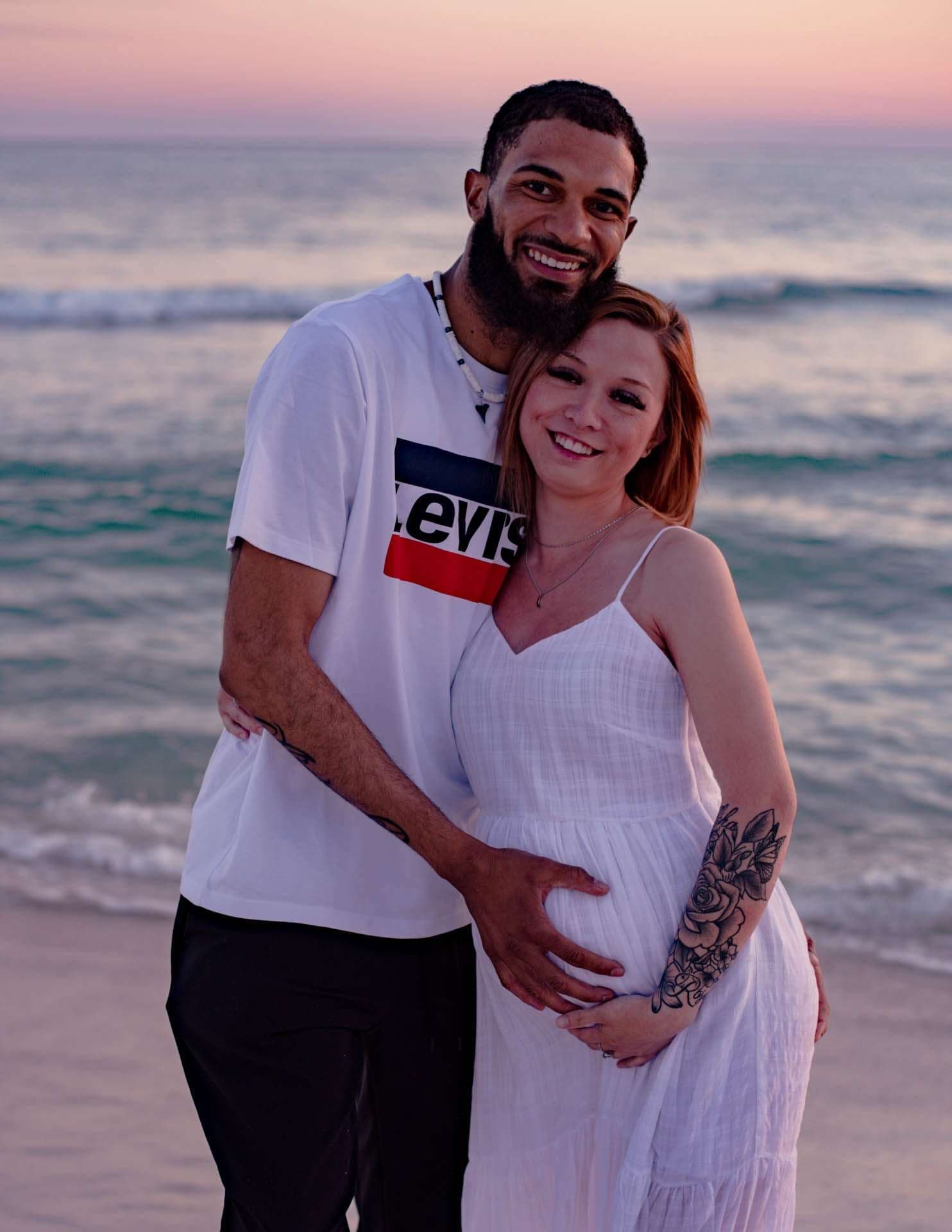 Smiling couple embracing on the beach at sunset, her hands on her baby bump, in white with the ocean behind