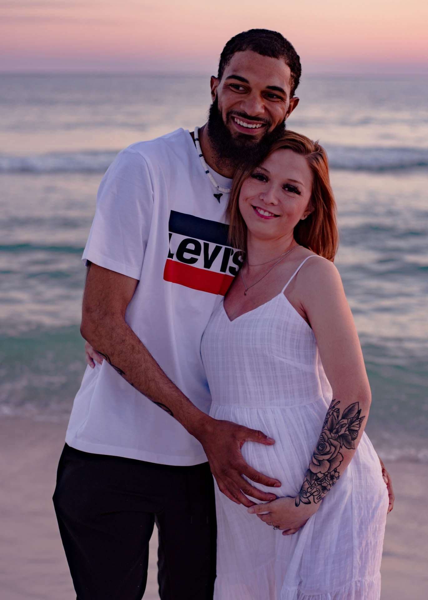 Smiling expectant couple embracing on the beach at sunset, his hand on her baby bump, she in a white dress
