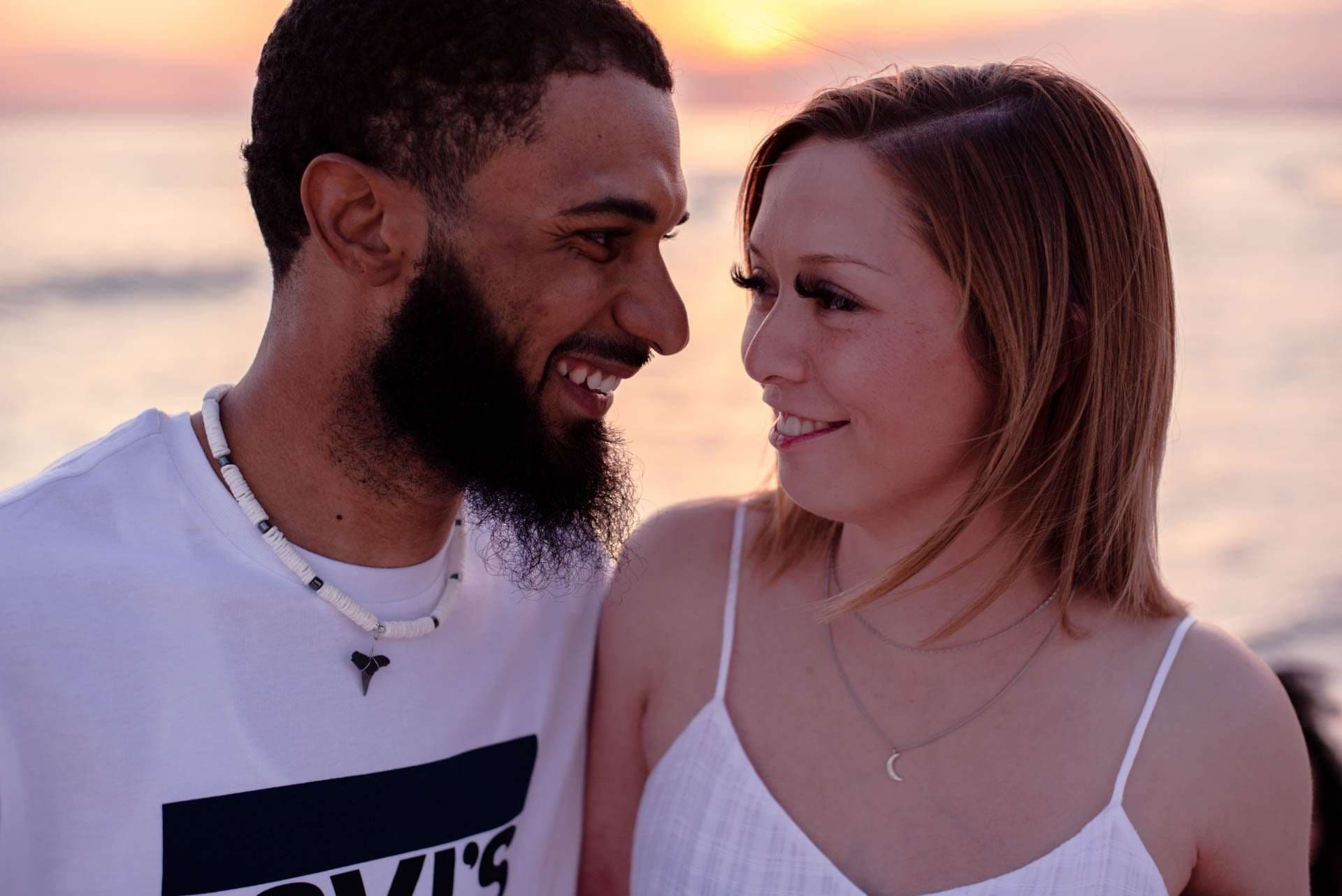 Smiling couple gazing at each other closely on the beach at golden hour, woman in white dress, man wearing shell necklace