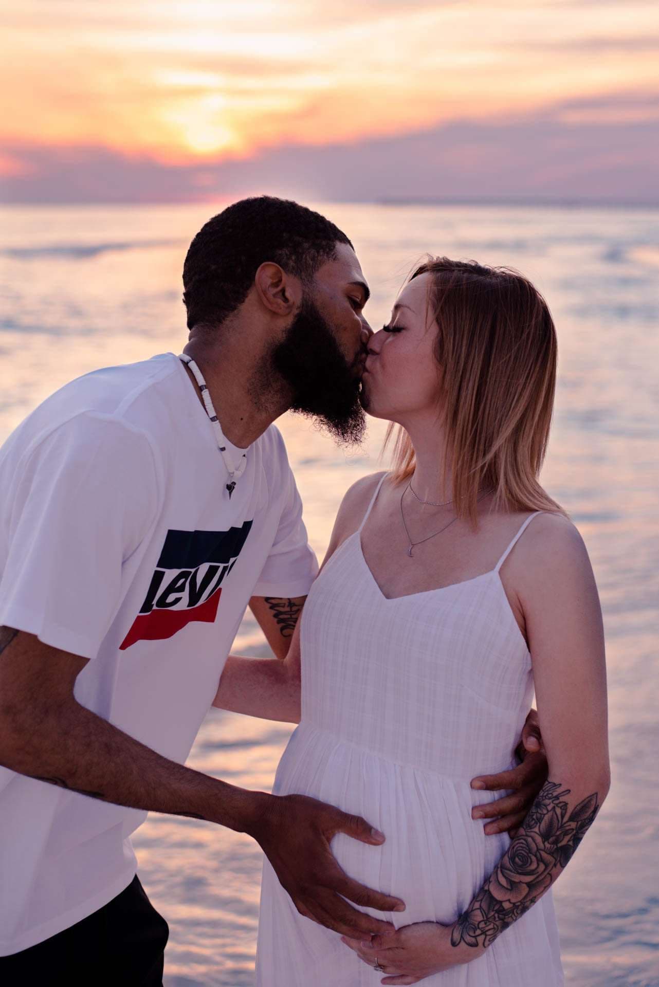 Couple kissing at the water's edge at sunset, hands resting on her pregnant belly, she in a white sundress