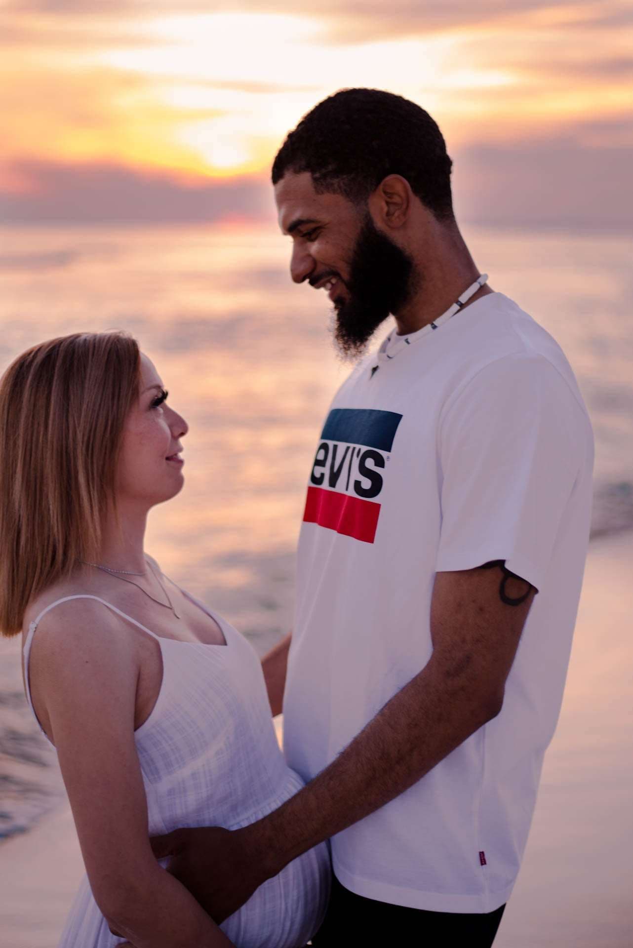 Couple smiling at each other on the beach at golden hour, woman in a white dress, man in a Levi's tee
