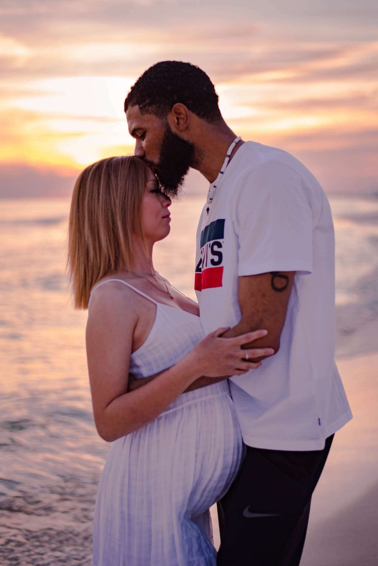 Bearded man kissing the forehead of his pregnant partner as they embrace on the beach at golden hour sunset