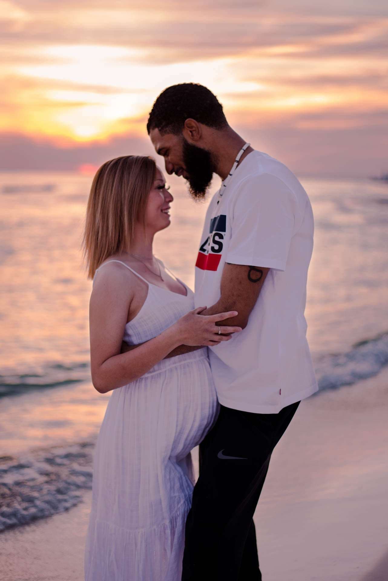 Expecting couple embracing forehead to forehead on the beach at golden hour, woman in a white dress