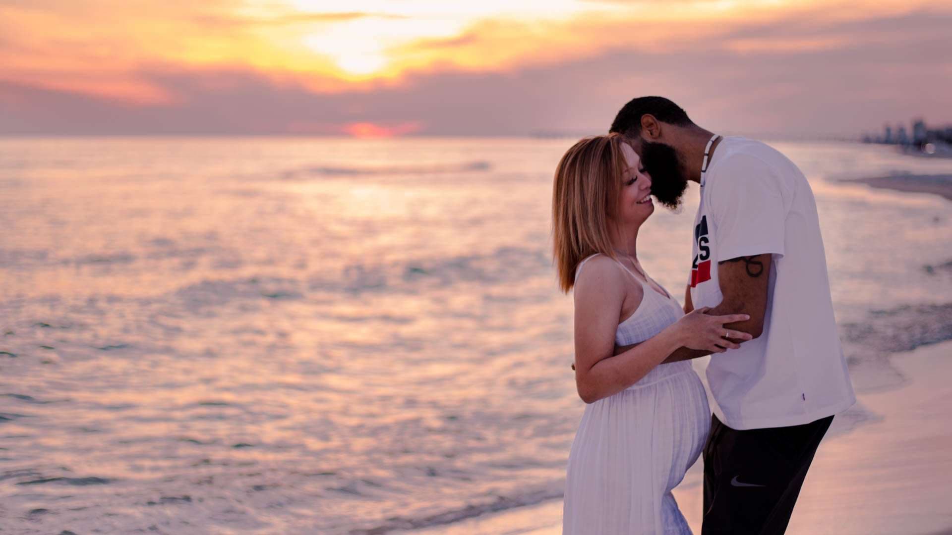 Expecting couple embracing on the beach at golden hour, the man nuzzling his partner's forehead in a white dress