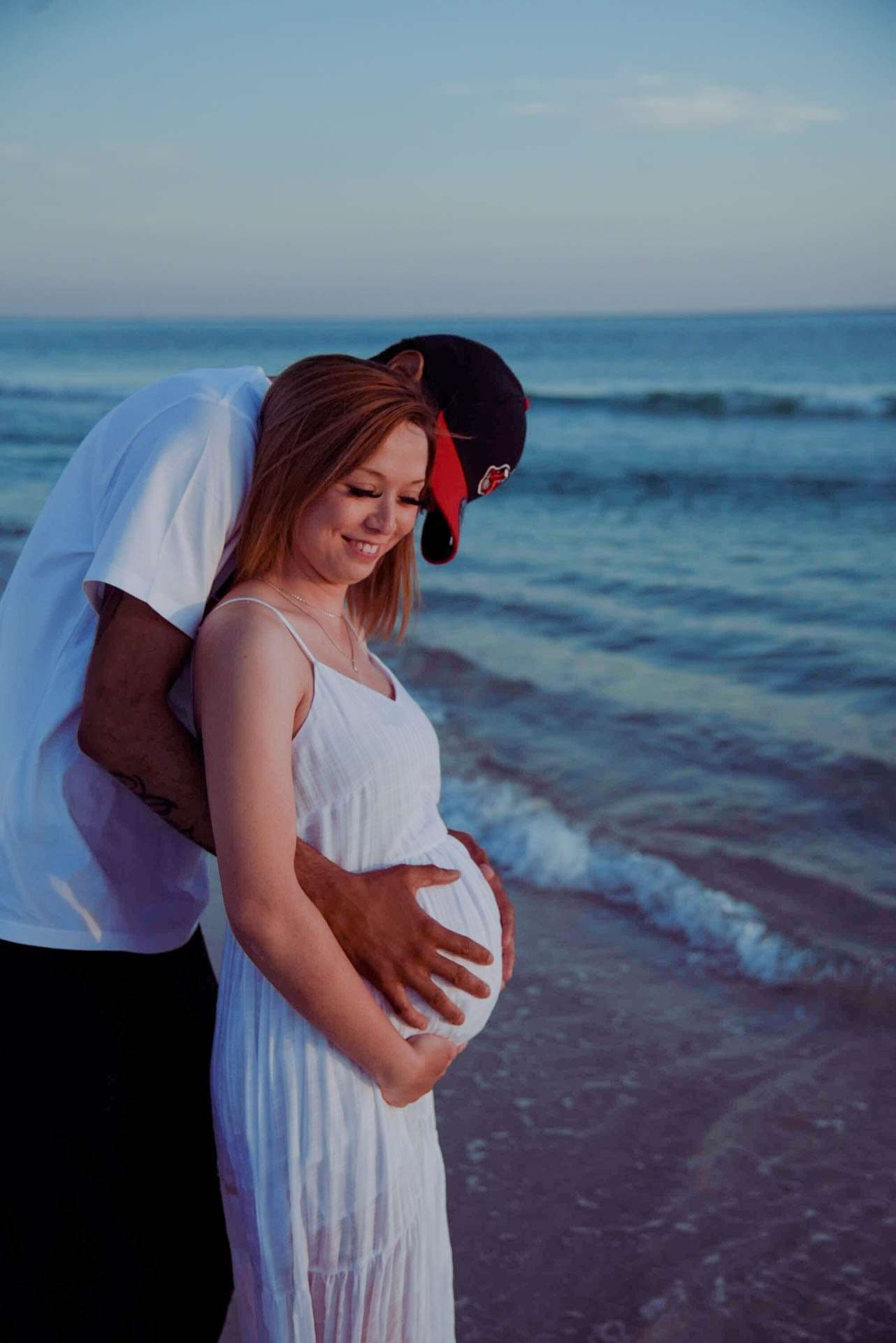 Expectant couple embracing on the beach at dusk, he kisses her head while holding her baby bump, she wears a white dress