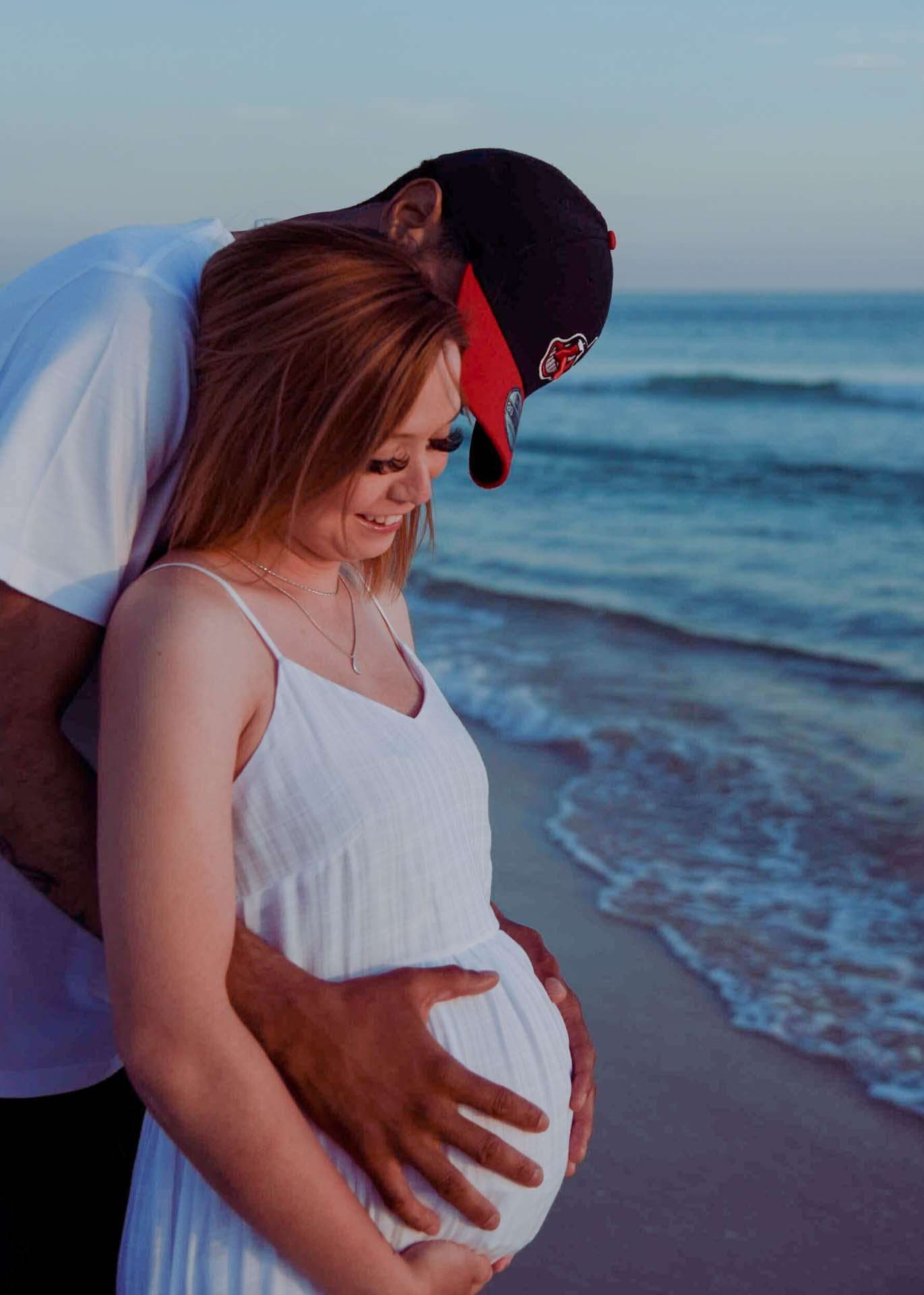Smiling pregnant woman in a white dress with a man in a cap cradling her baby bump by the ocean at dusk