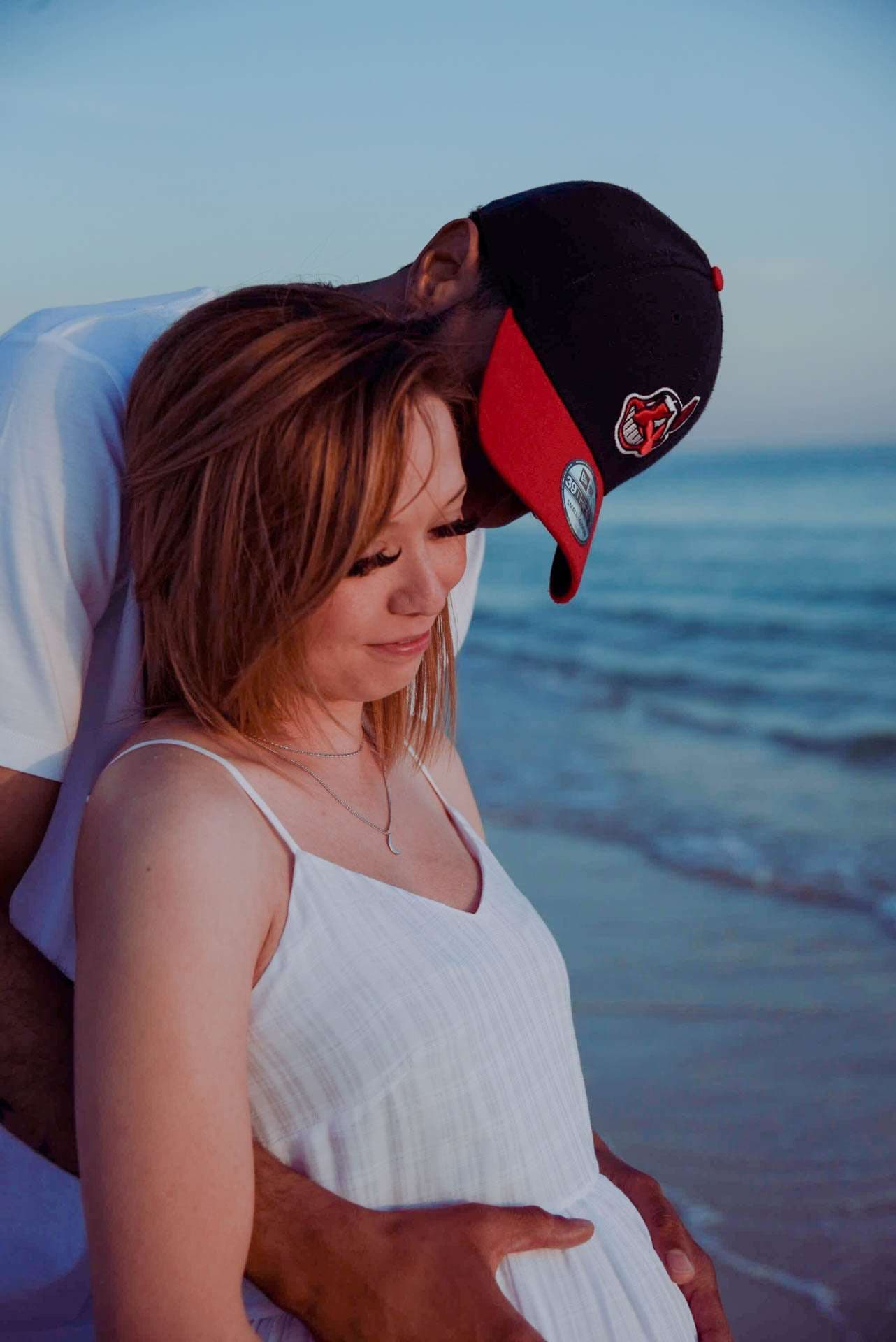 Man in a black cap leaning down to kiss a woman in a white sundress, his hand on her belly, by the ocean
