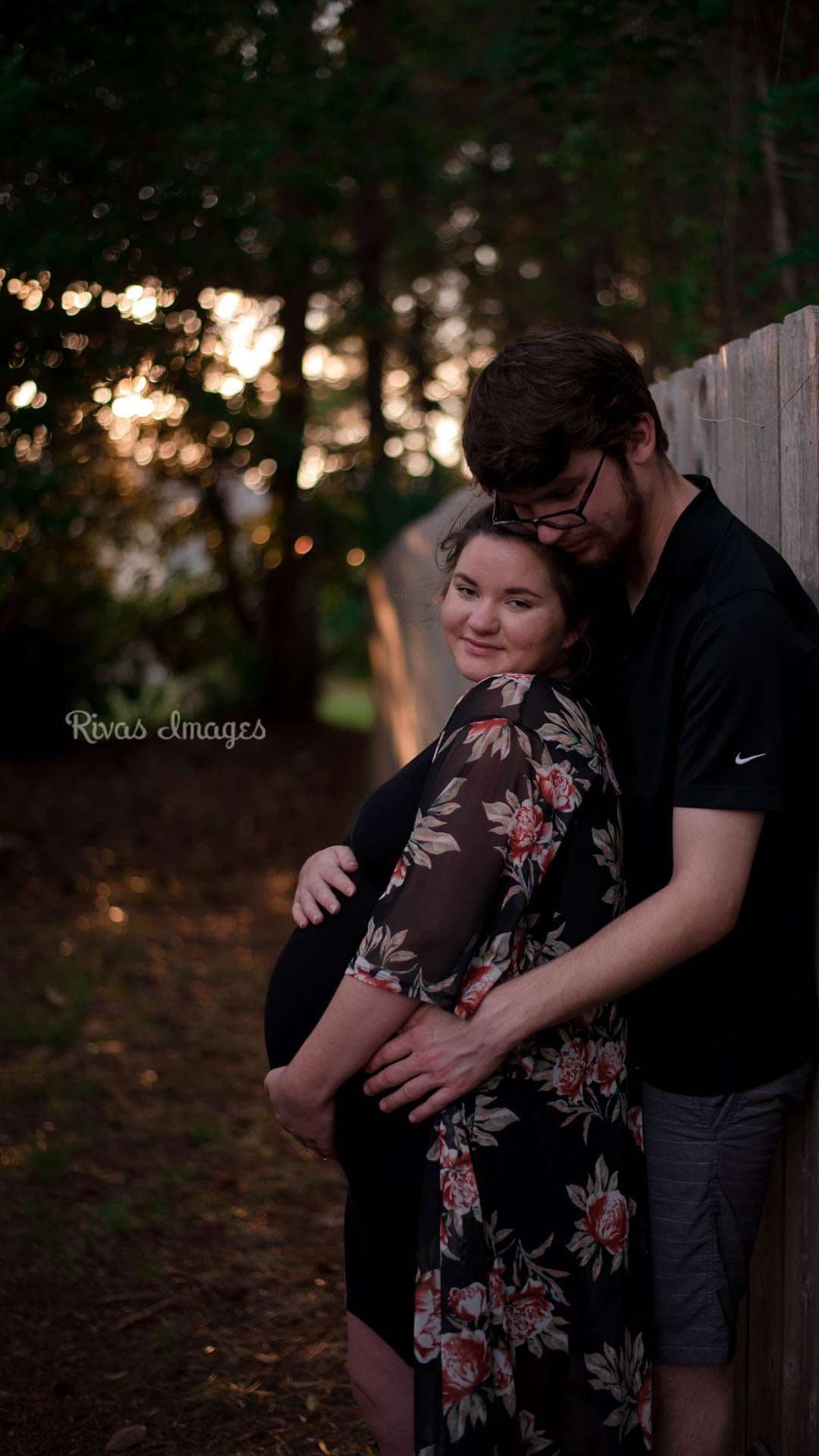 Man embracing a pregnant woman in a floral dress from behind beside a wooden fence in the woods at sunset