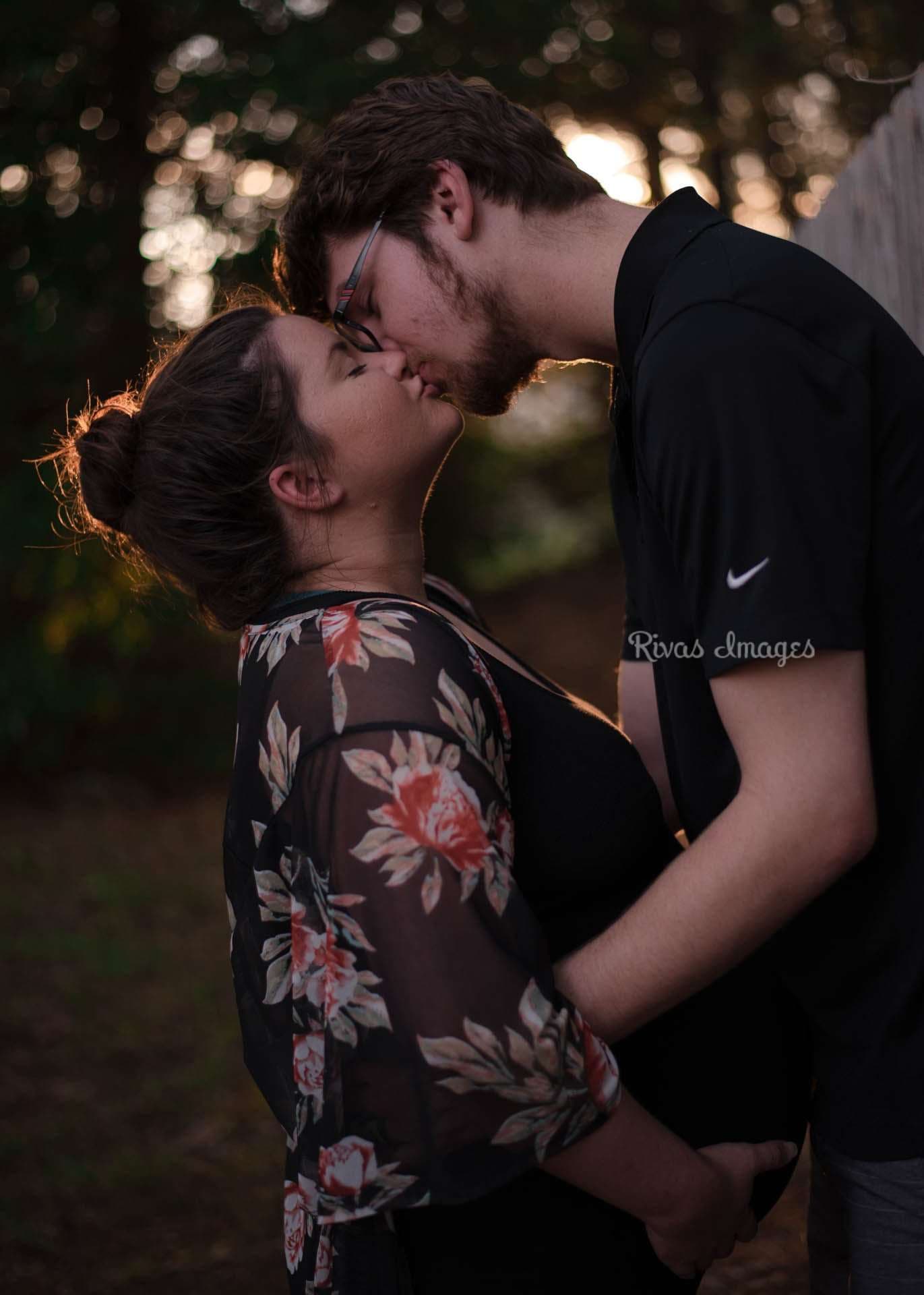 Couple kissing outdoors at sunset among trees, her floral kimono over a black dress, his hand on her belly