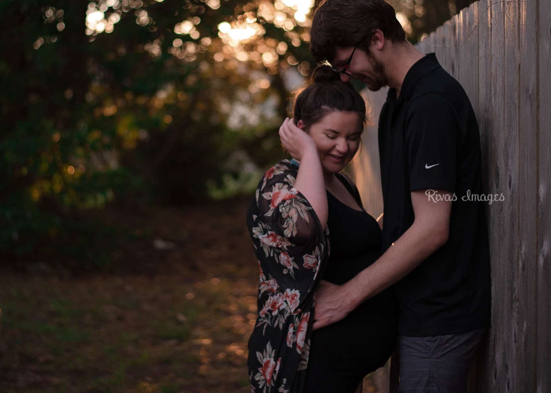 Smiling expectant woman in a floral kimono leans into her partner beside a wooden fence at golden sunset